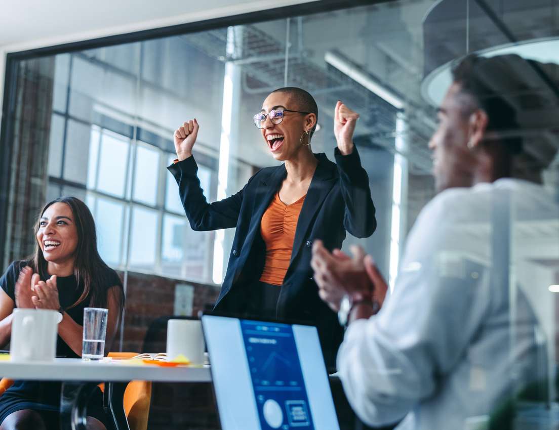 A diverse group of colleagues celebrating in an office meeting. A woman with short hair and glasses raises her arms in triump