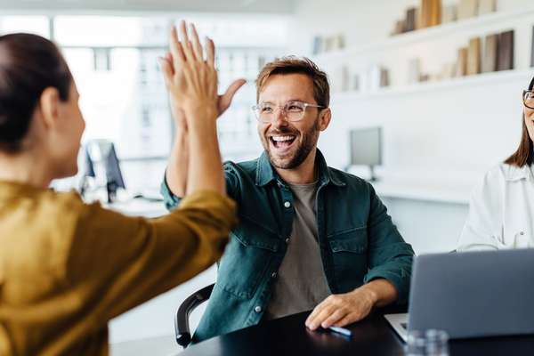Two colleagues high-fiving in a bright office, a man laughing joyfully, while a woman looks on smiling.