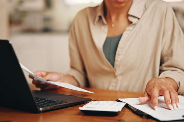Person calculating finances, holding papers, and using a laptop and calculator on a wooden desk. Wearing a light brown shirt.
