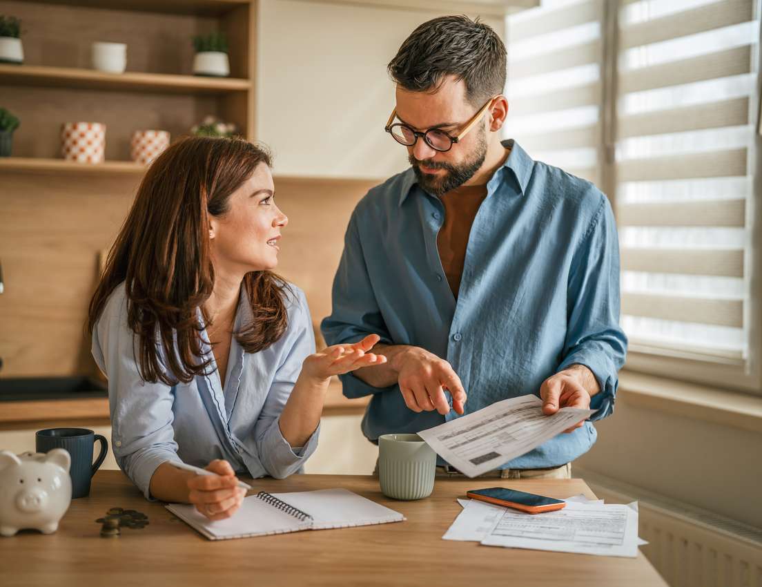 A couple in a kitchen discusses household finances. The man holds bills, the woman explains while writing in a notebook, with