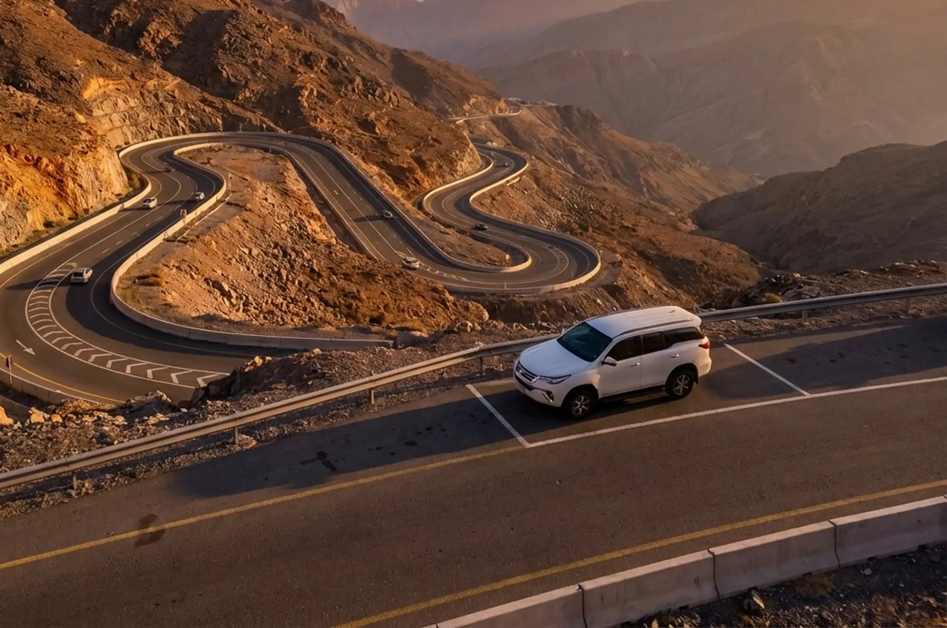 White SUV parked by a scenic, winding mountain road at sunset with rugged terrain and warm hues in the background.