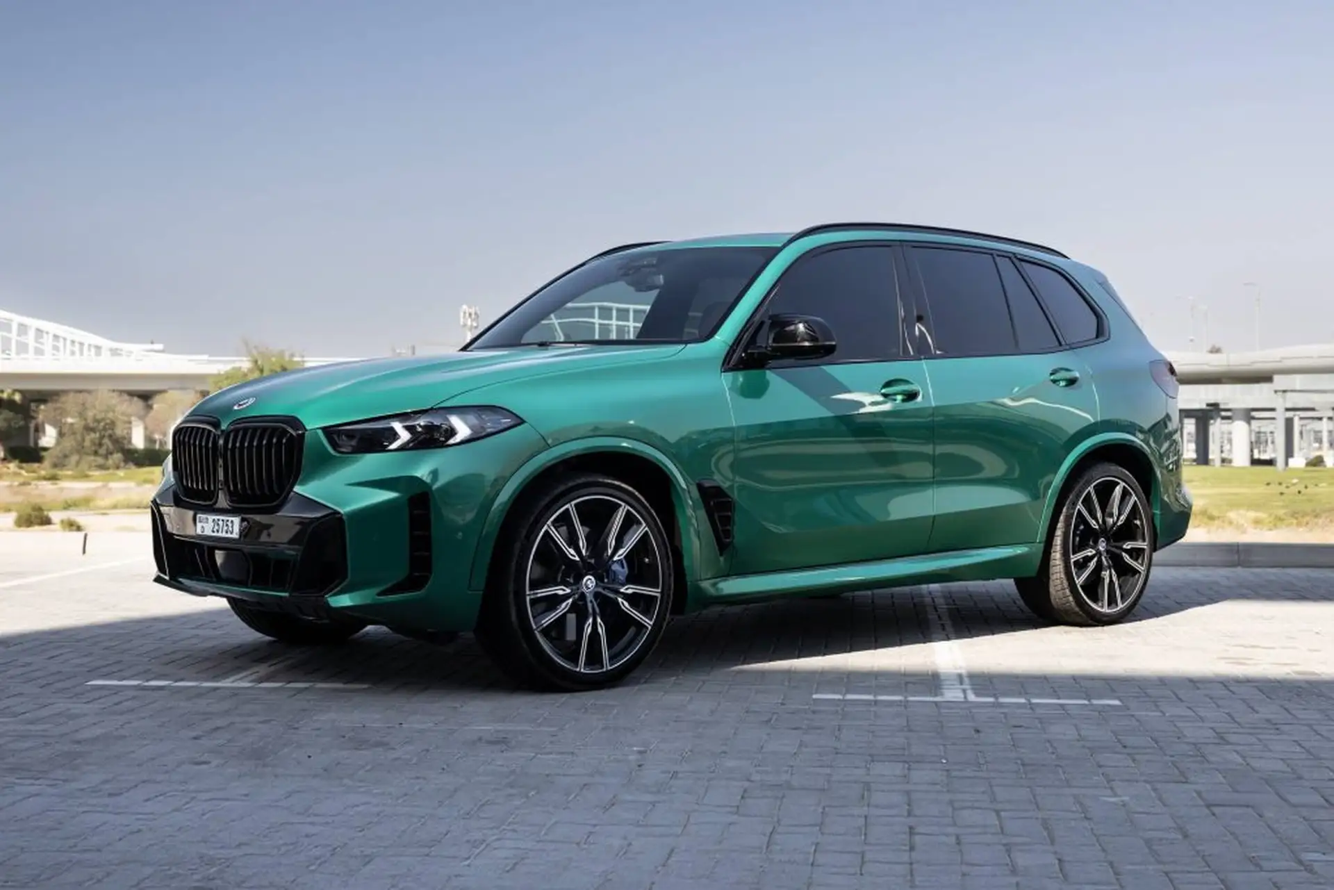 Green BMW SUV parked on a paved area, featuring sleek black rims and tinted windows, with a clear sky and modern architecture in the background.