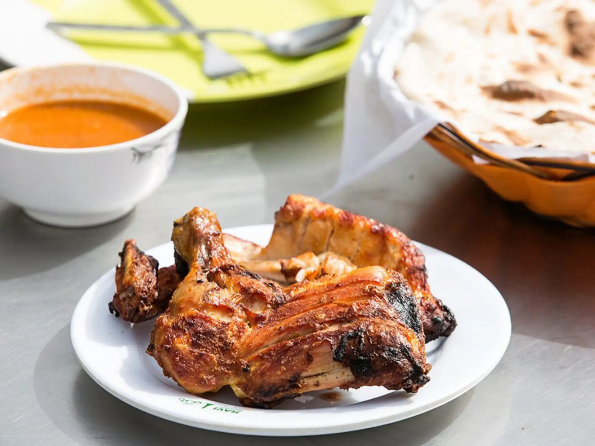 Grilled chicken on a white plate, accompanied by a bowl of red sauce, naan bread in a basket, with utensils on a table.