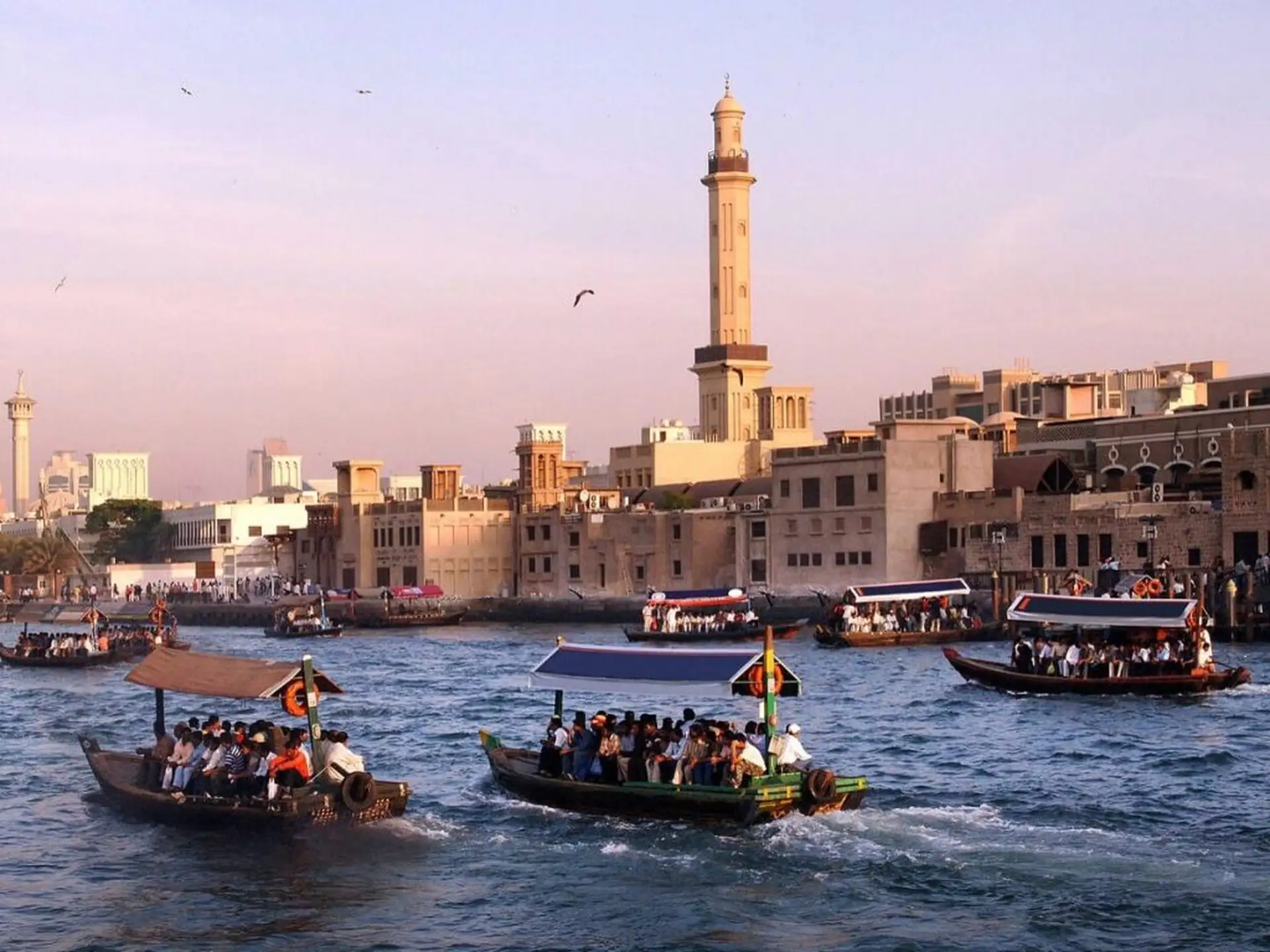 Boats filled with passengers traverse a bustling waterway in front of Dubai's traditional buildings and a tall minaret under a clear sky.
