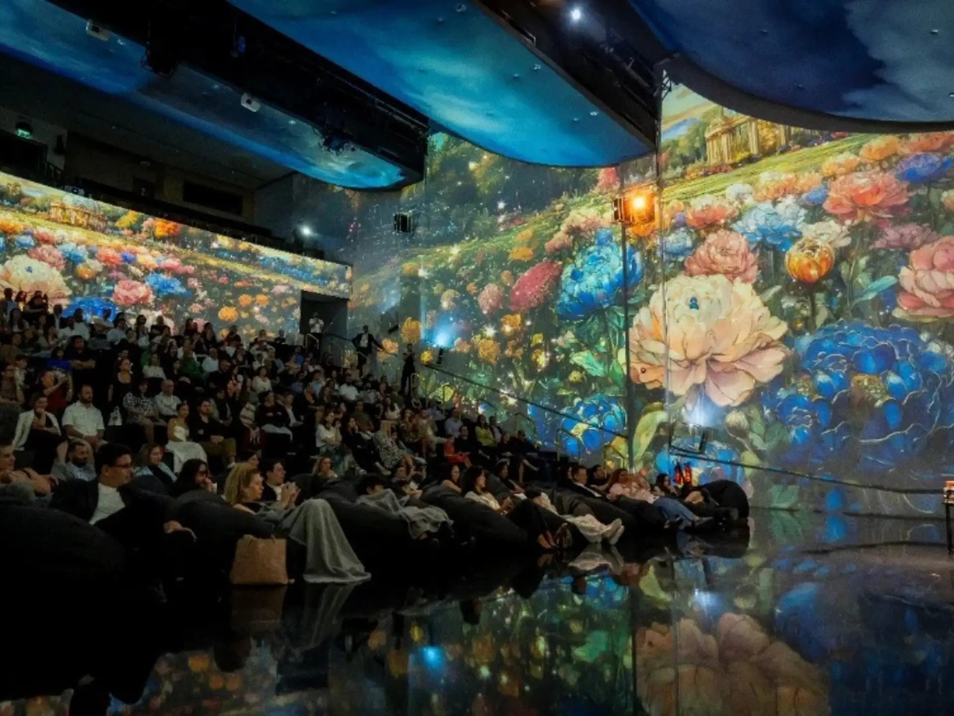 Audience watching vibrant floral projections on walls and ceiling in a dark auditorium. The atmosphere is immersive and artistic.