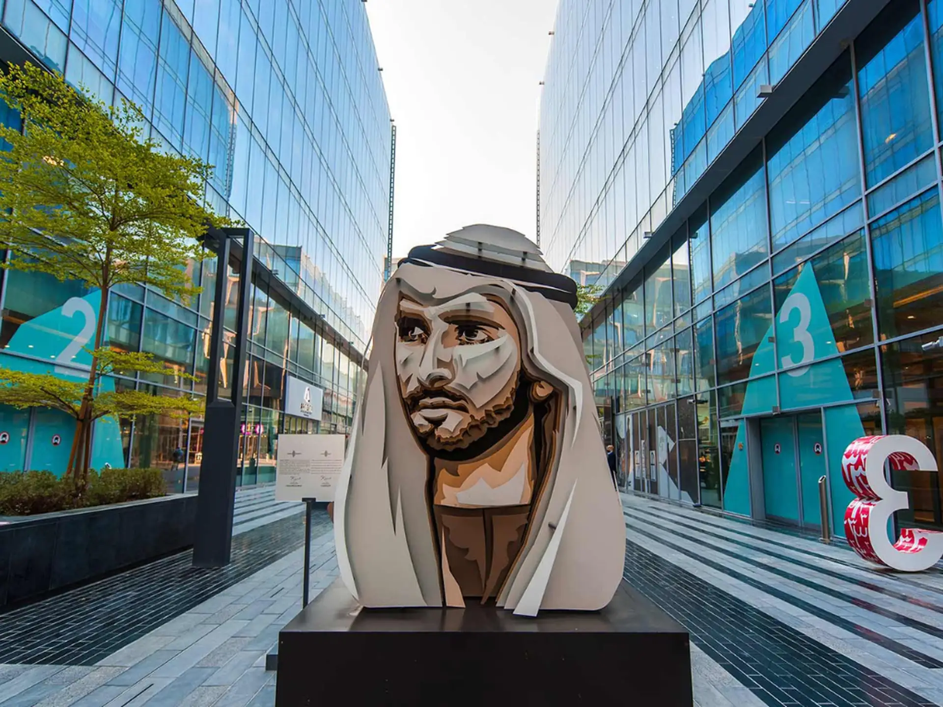 Bust sculpture of a man in traditional attire, set between modern glass buildings with numbers "2" and "3" visible, and greenery nearby.