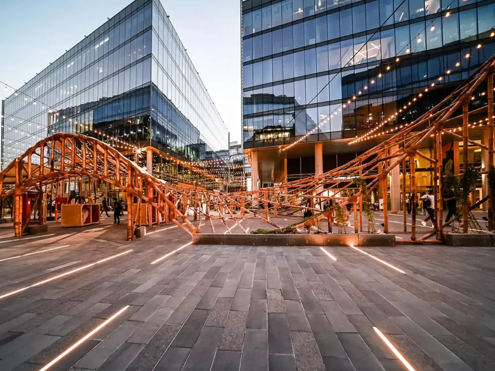 Modern plaza with tall glass buildings and an intricate wooden installation, adorned with string lights, at dusk.
