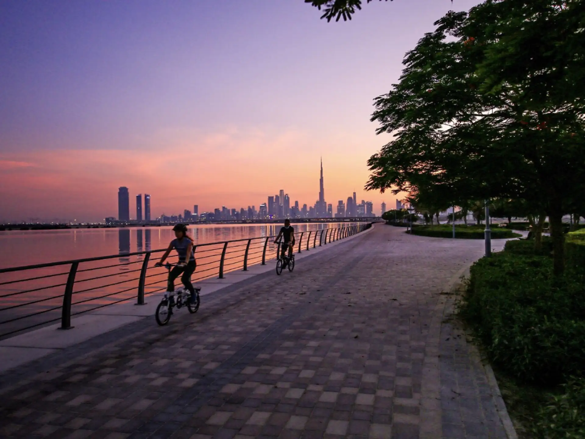 Two cyclists ride along a waterfront path at sunset, with a city skyline featuring a tall skyscraper in the background.