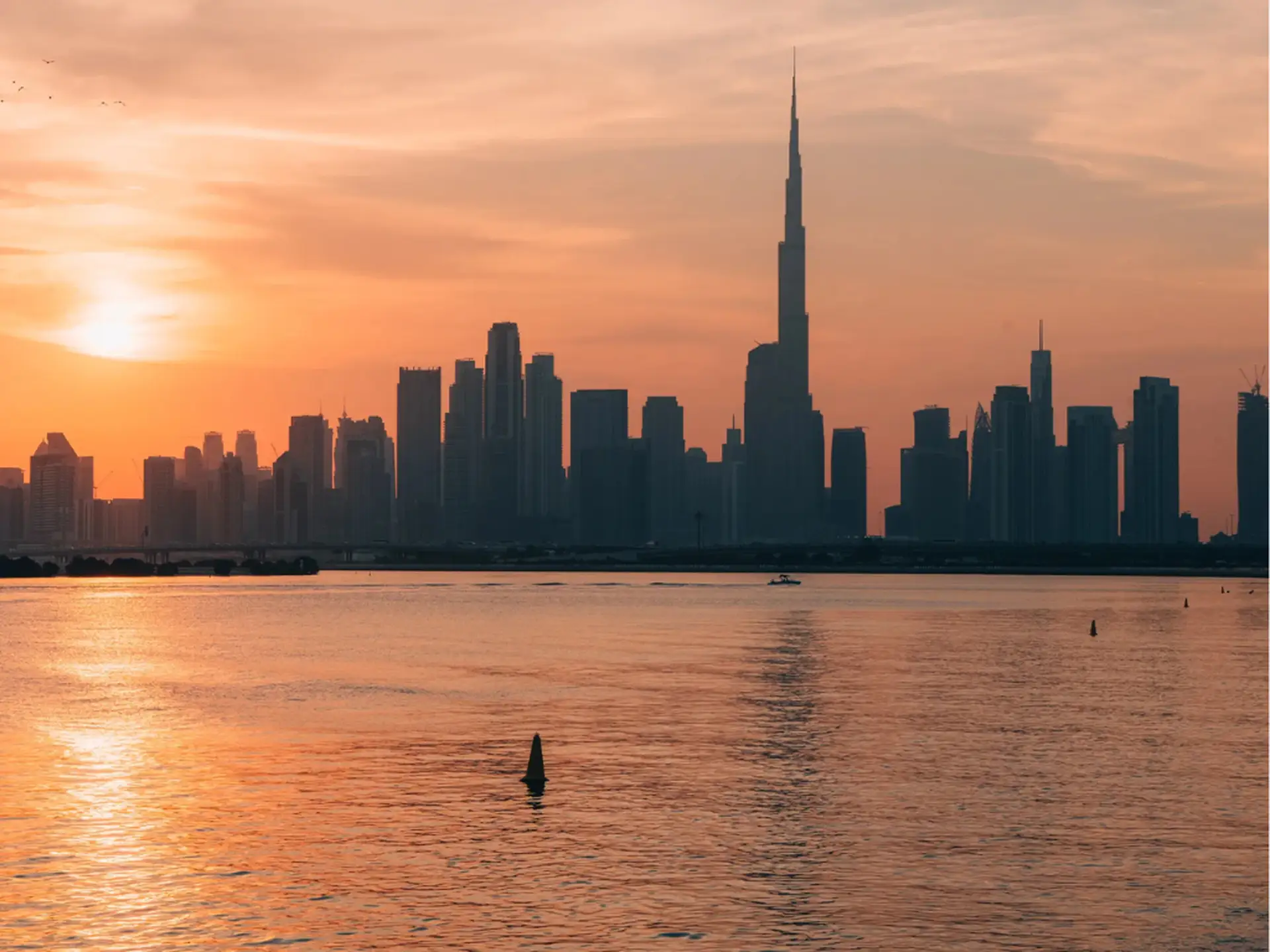 Skyline of a city with a tall spire under an orange sunset, reflecting on calm waters in the foreground.