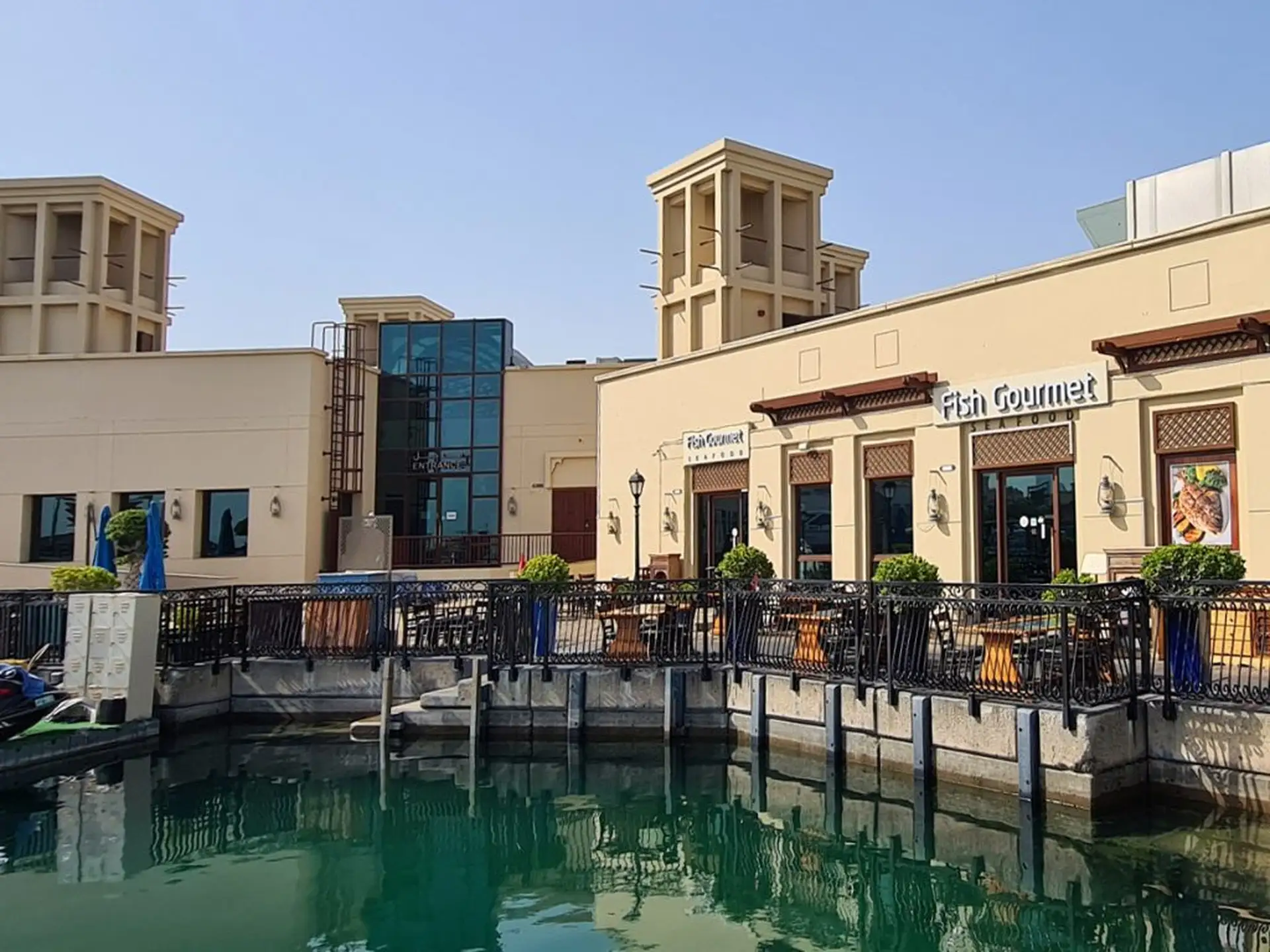 Waterfront dining area with outdoor seating, surrounded by buildings and clear waters under a blue sky.