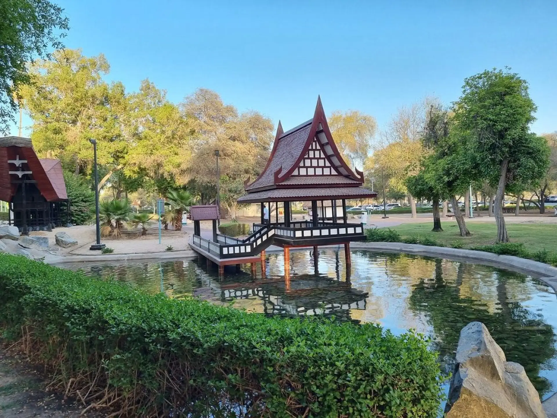 A traditional-style pavilion with a steep roof stands over a reflective pond, surrounded by lush greenery and trees in a park setting.