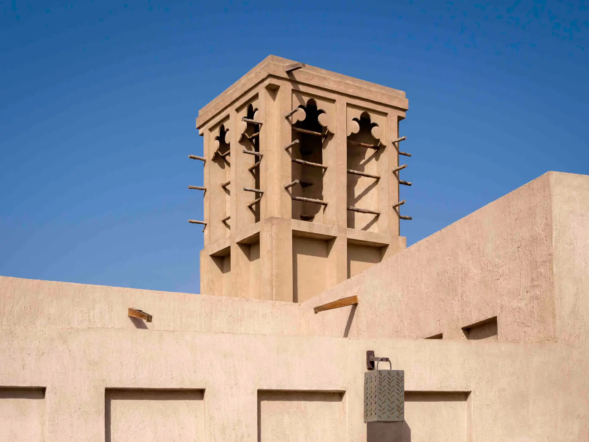 A beige wind tower with wooden beams and intricate cutouts against a clear blue sky, part of a traditional Middle Eastern-style building.