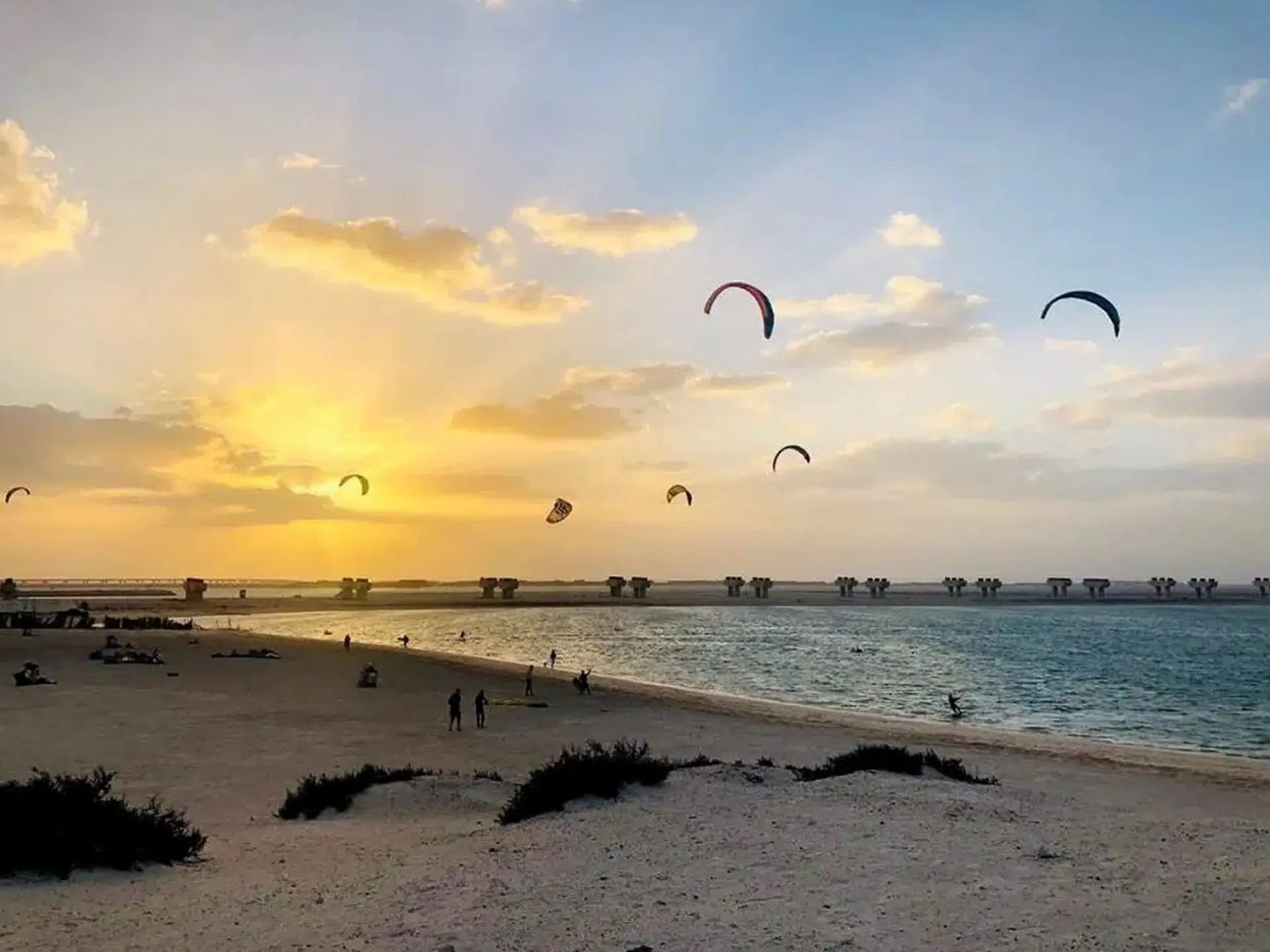 Kitesurfers glide over the ocean at sunset, with a sandy beach and silhouetted figures in the foreground, and scattered clouds in the sky.
