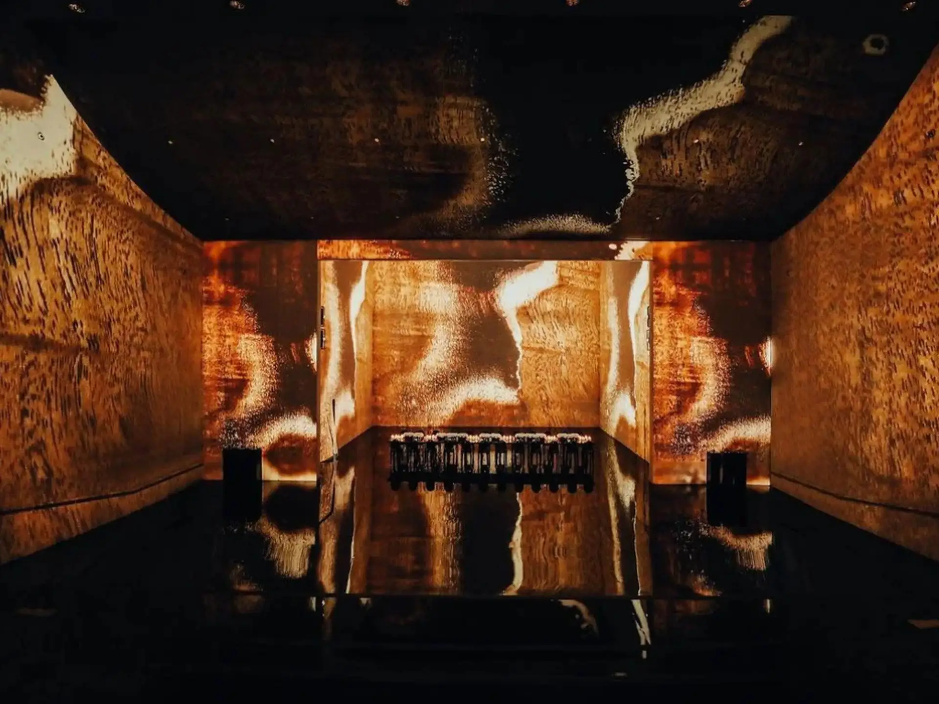 A dimly lit room with abstract brown and gold projections on walls and ceiling, reflecting on a glossy floor with a central display.
