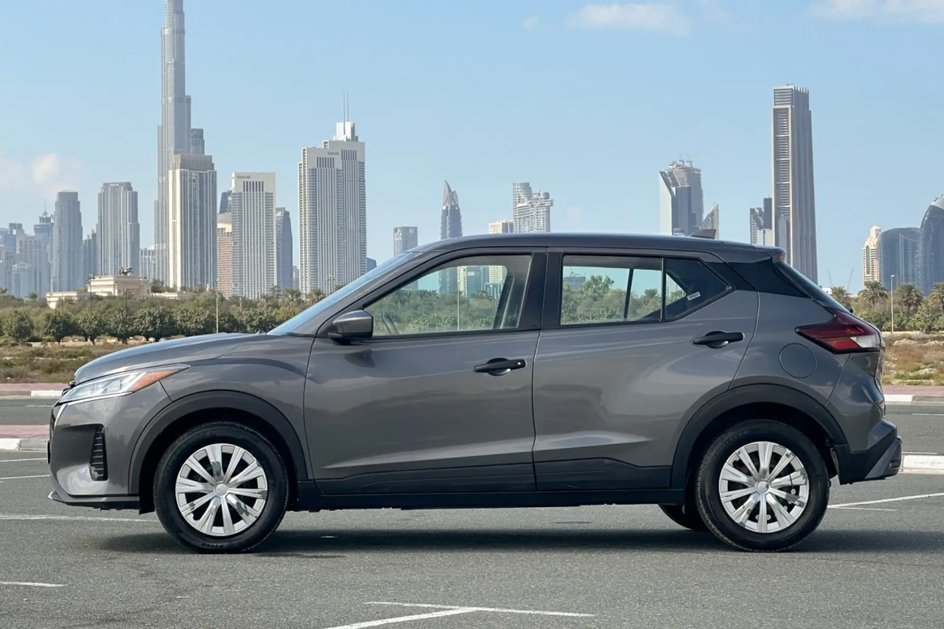A gray SUV is parked on a road with a city skyline featuring modern skyscrapers in the background under a clear blue sky.
