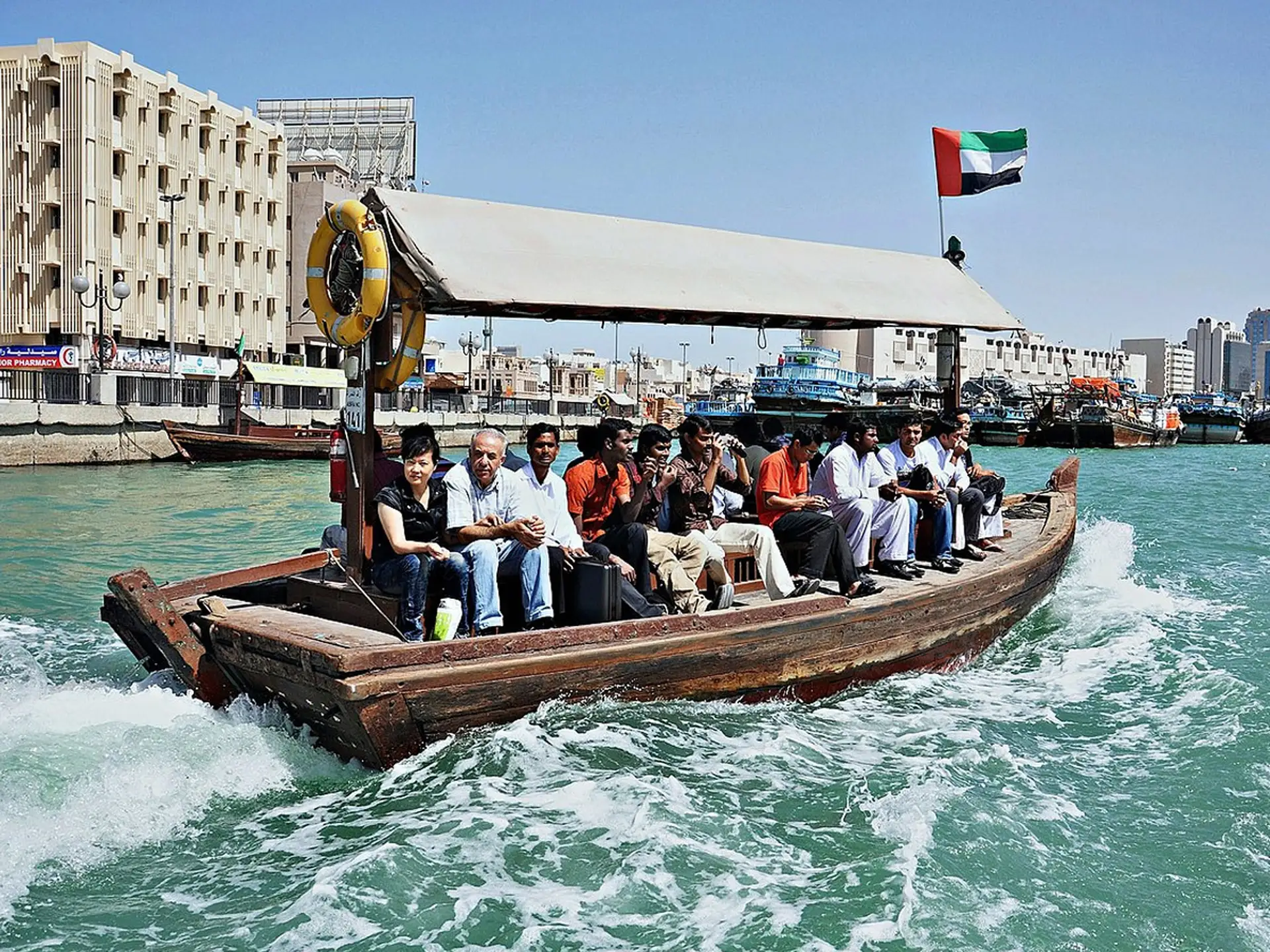 A wooden boat carrying passengers on a turquoise waterway, with an Emirati flag flying, against a backdrop of buildings and clear blue sky.