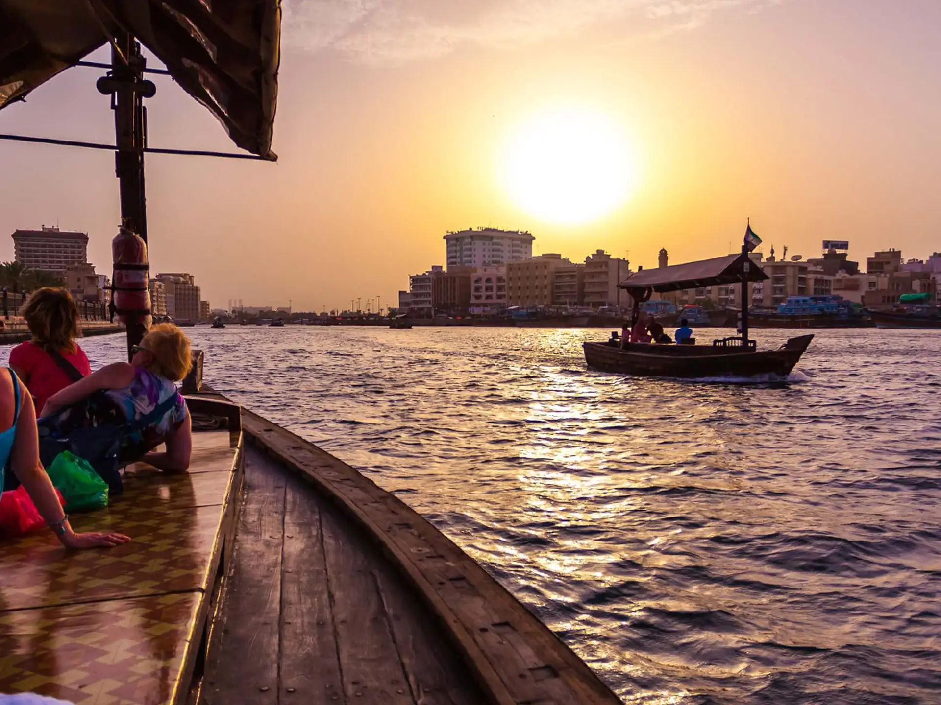 Sunset view from a wooden boat on a busy river, with city buildings silhouetted in the background and another boat passing by.