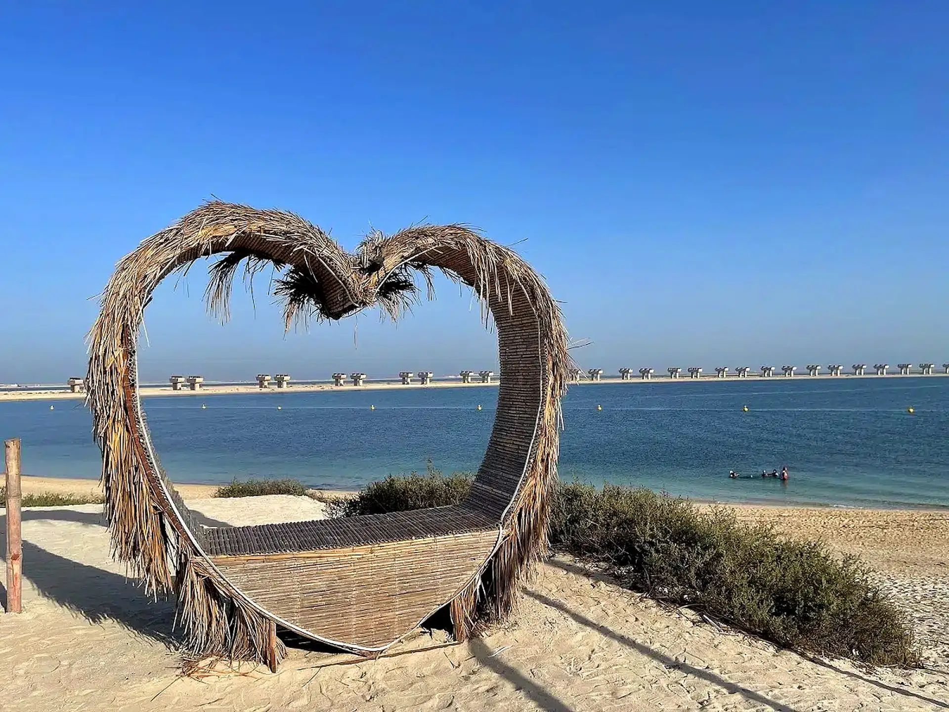 Heart-shaped palm-frond bench on a sandy beach overlooking calm blue water and a distant bridge under a clear blue sky.