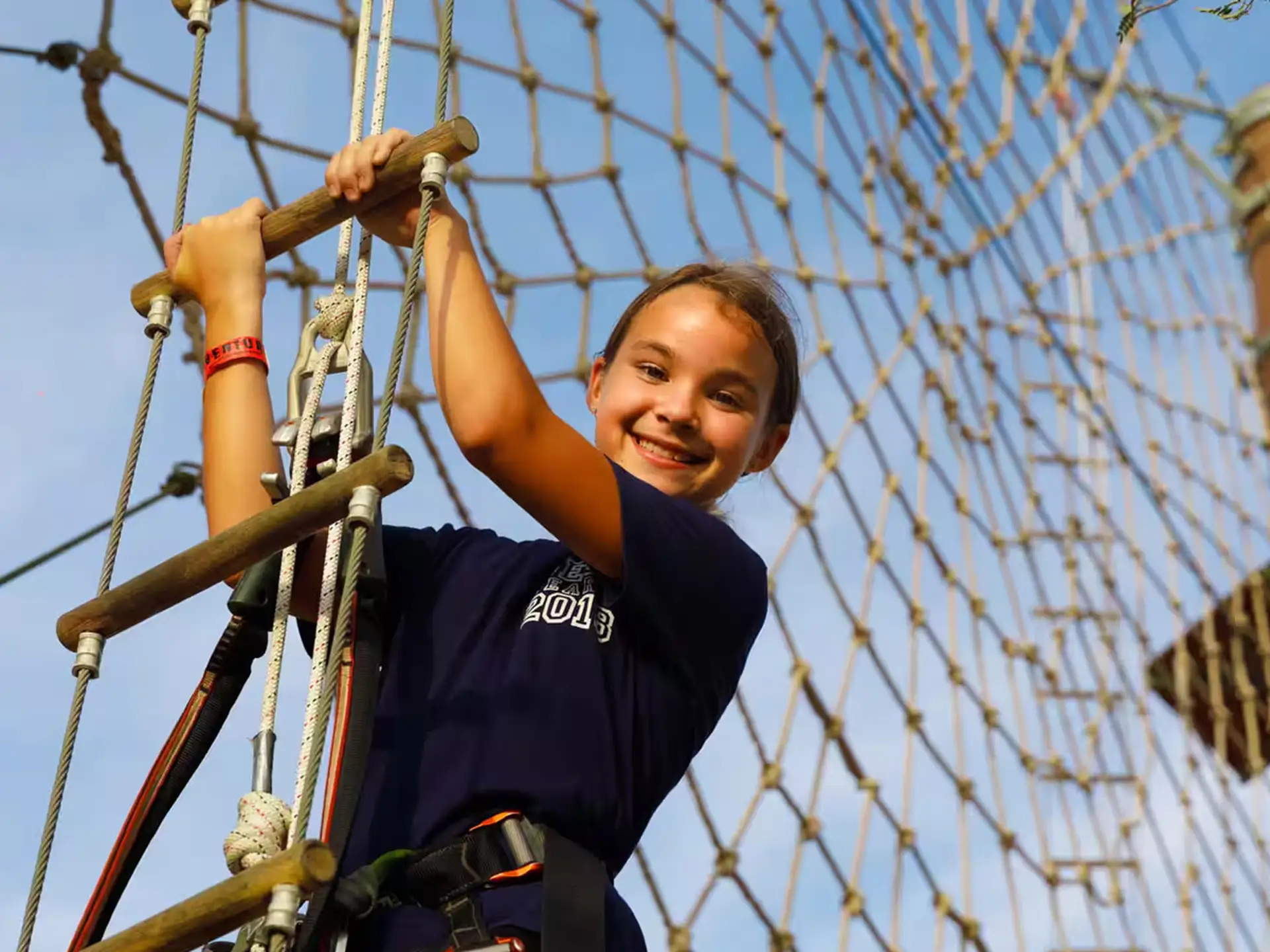 A smiling girl climbs a rope course on a sunny day, wearing a harness and gripping the ropes confidently.