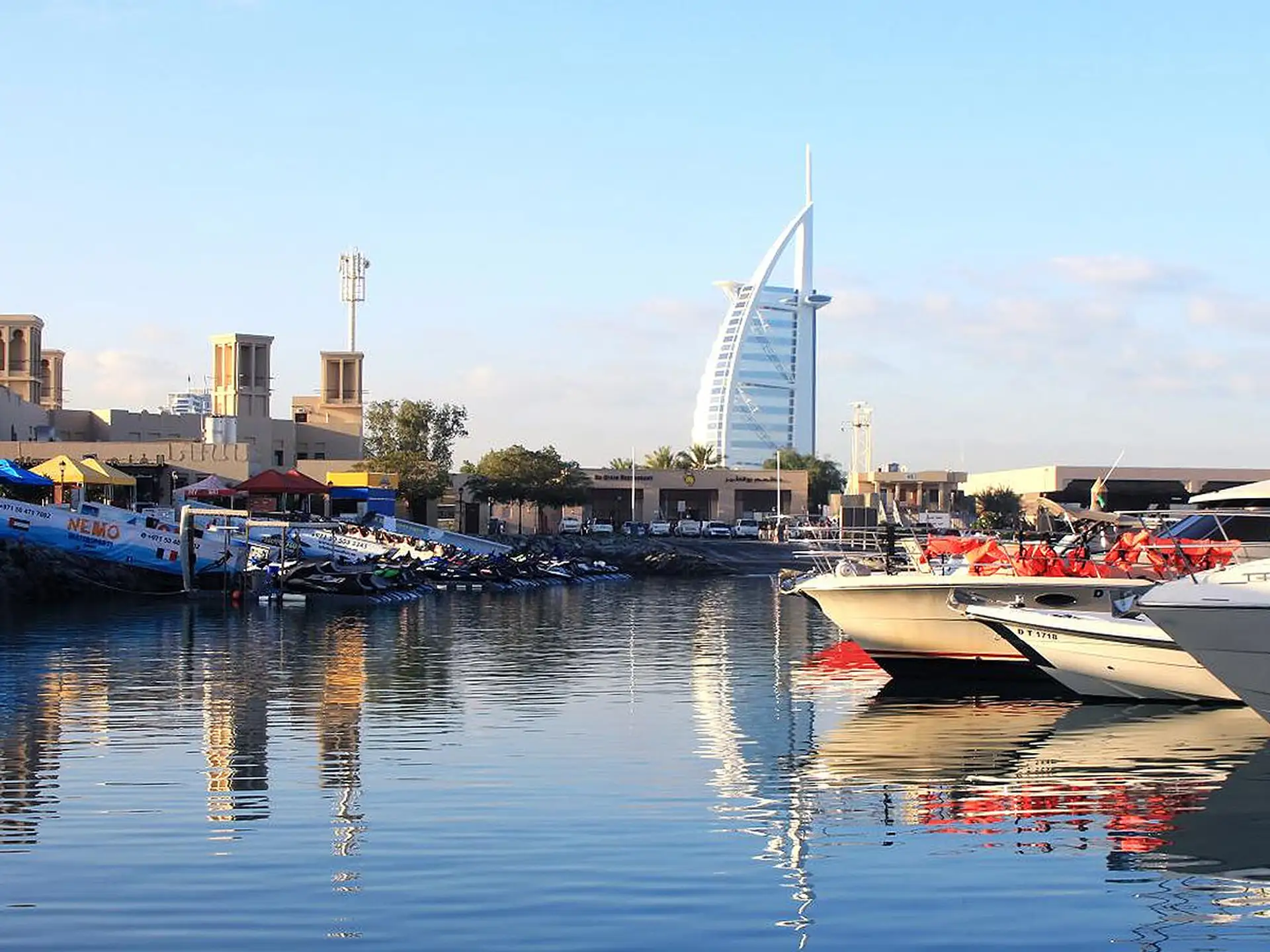 Boats docked in a marina with calm water, modern buildings in the background, and a distinctive sail-shaped skyscraper under a clear sky.