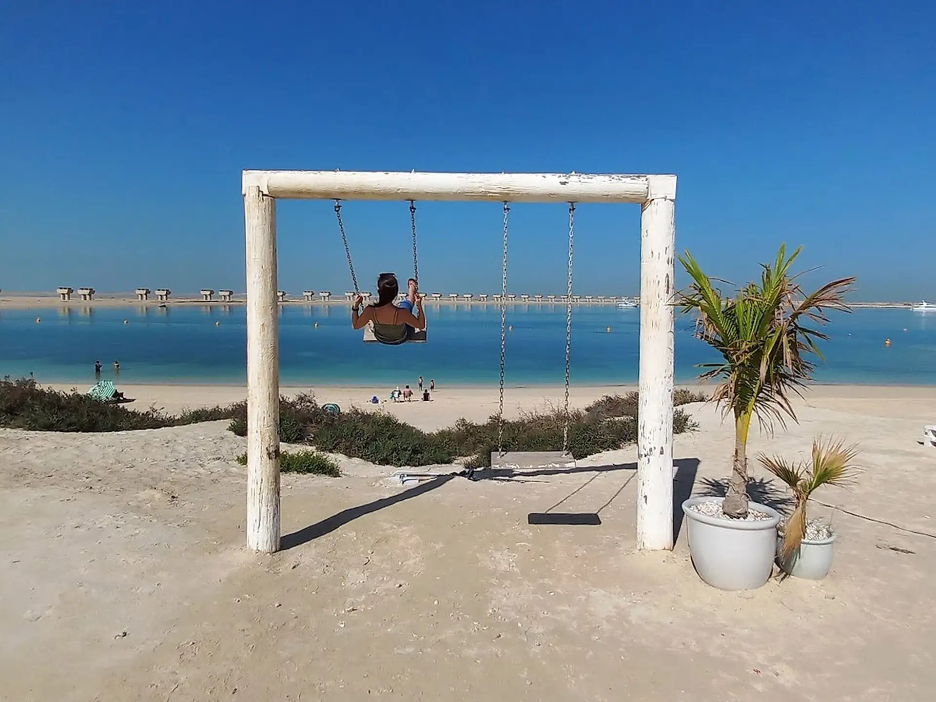 Person swinging on a beach swing facing a calm, blue sea under a clear sky, with potted palm and beachgoers in the background.
