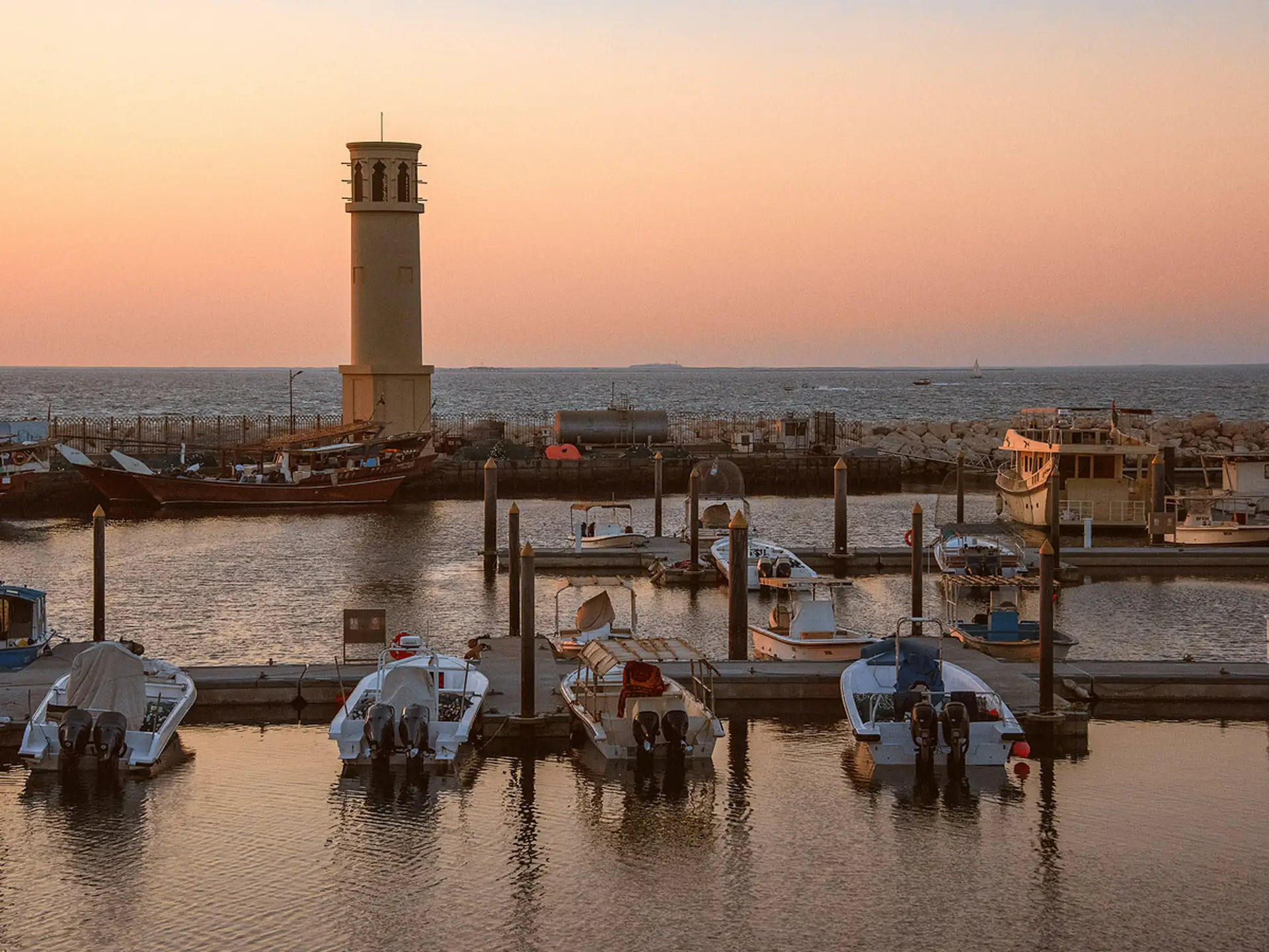Boats docked at a marina during sunset, with a lighthouse silhouetted against a pink and orange sky. Calm waters reflect the boats and sky.