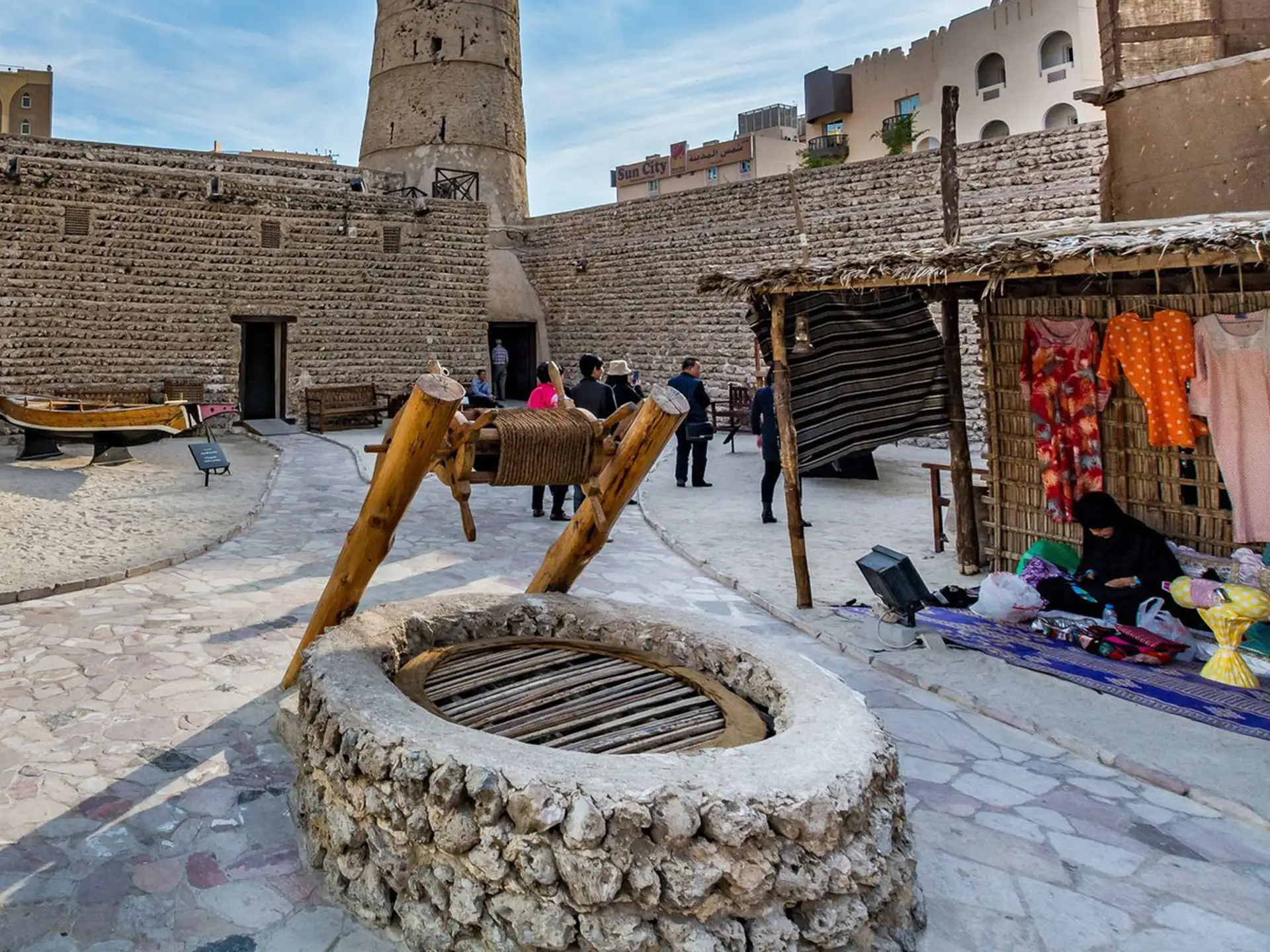 A historical courtyard with a stone well, traditional market stalls, and people exploring the area surrounded by ancient architecture.