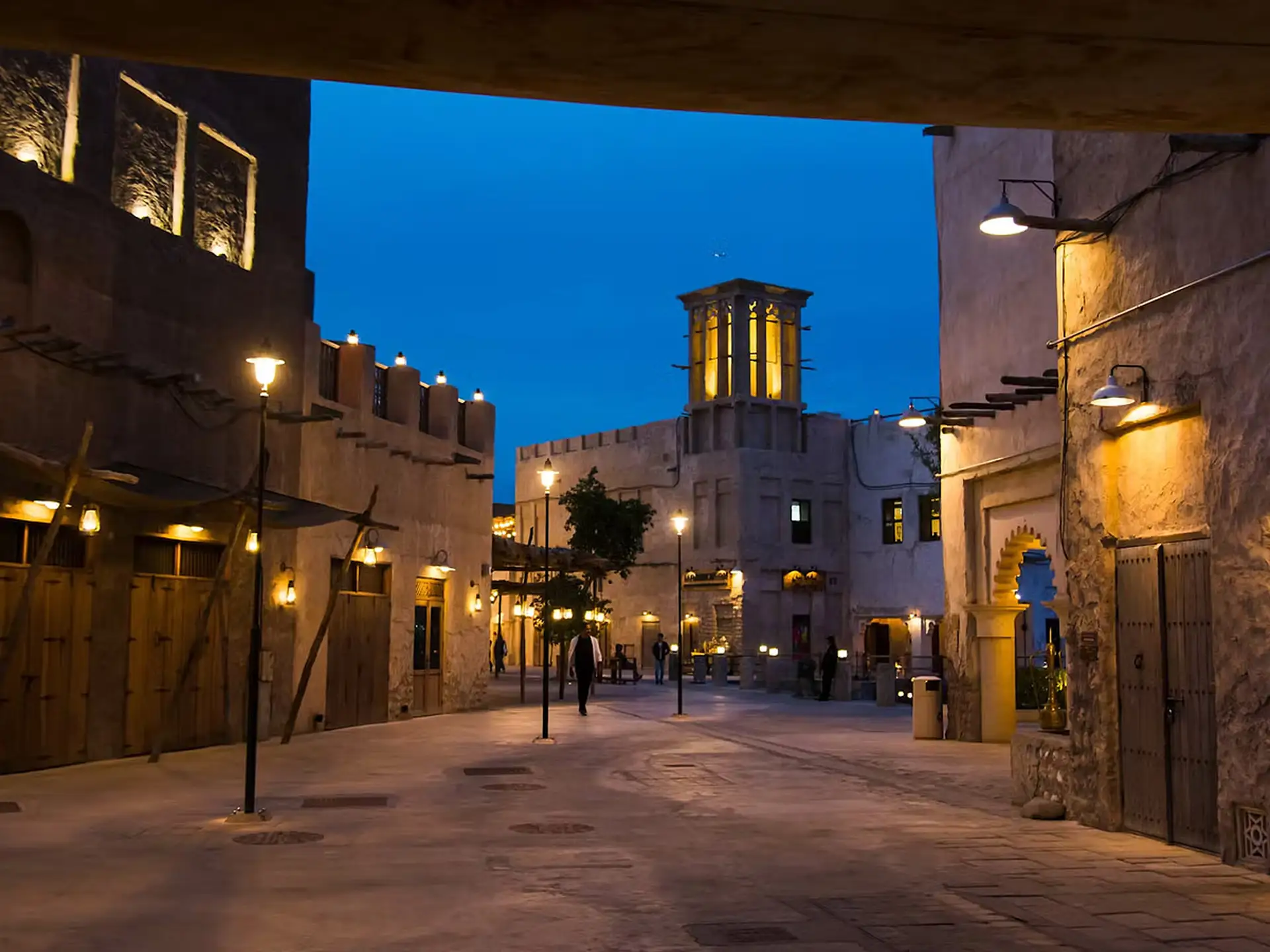 Night view of a beautifully lit traditional Middle Eastern street with stone buildings, lanterns, and a wind tower under a deep blue sky.