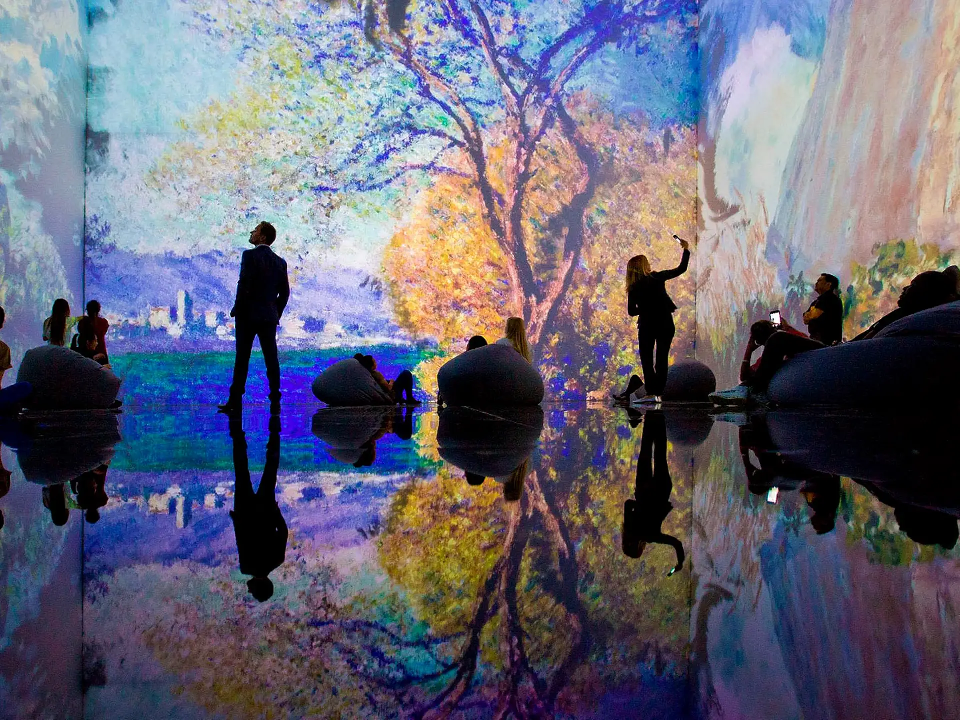 People in an immersive art exhibit with colorful digital projections of landscapes and reflections on a glossy floor.