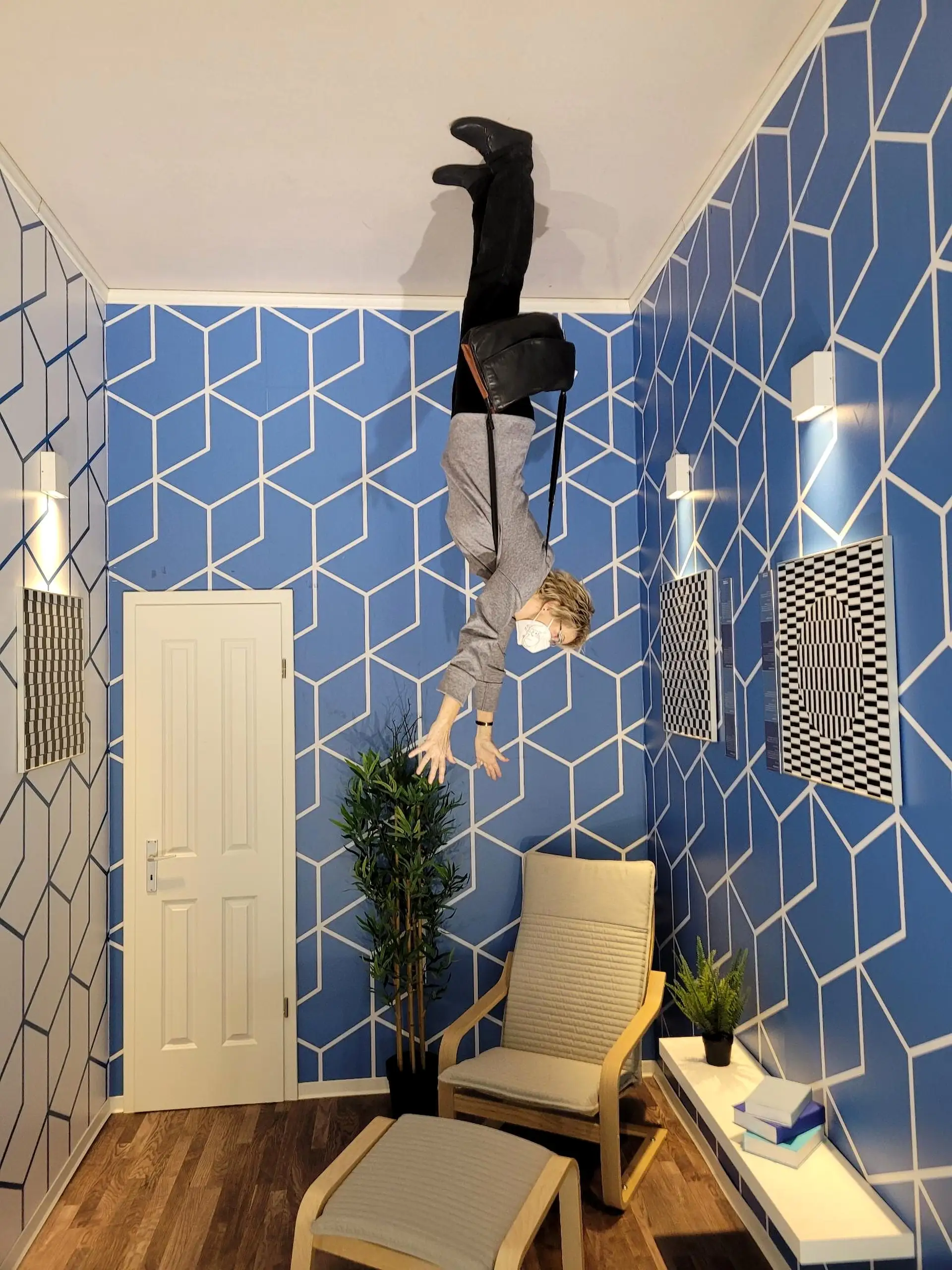 Person hanging upside down from the ceiling in a room with blue geometric wallpaper, a plant, and a chair below. Optical illusion effect.