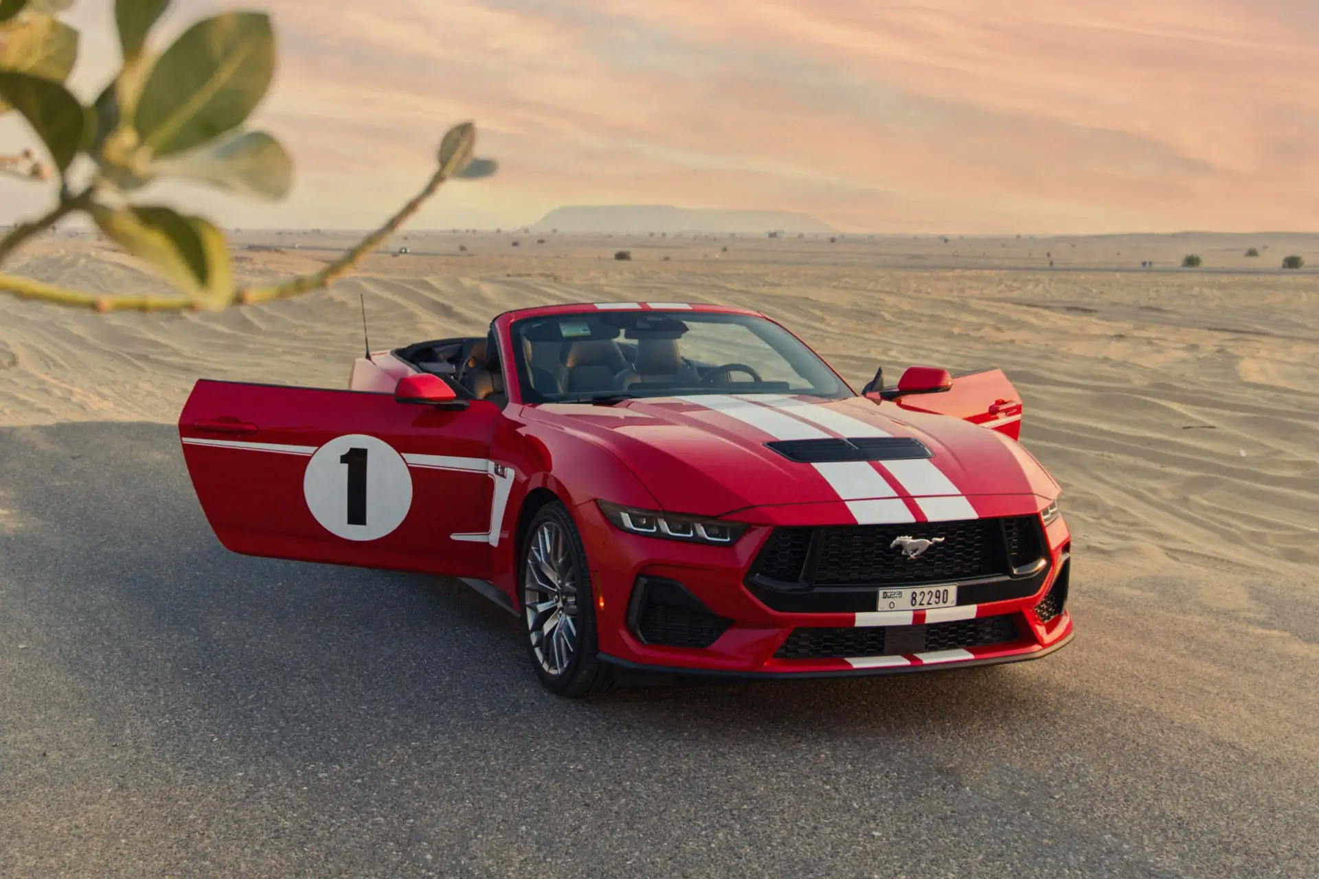 Red convertible sports car with white racing stripes and number 1 on the door, parked on a desert road at sunset. Open driver's door.