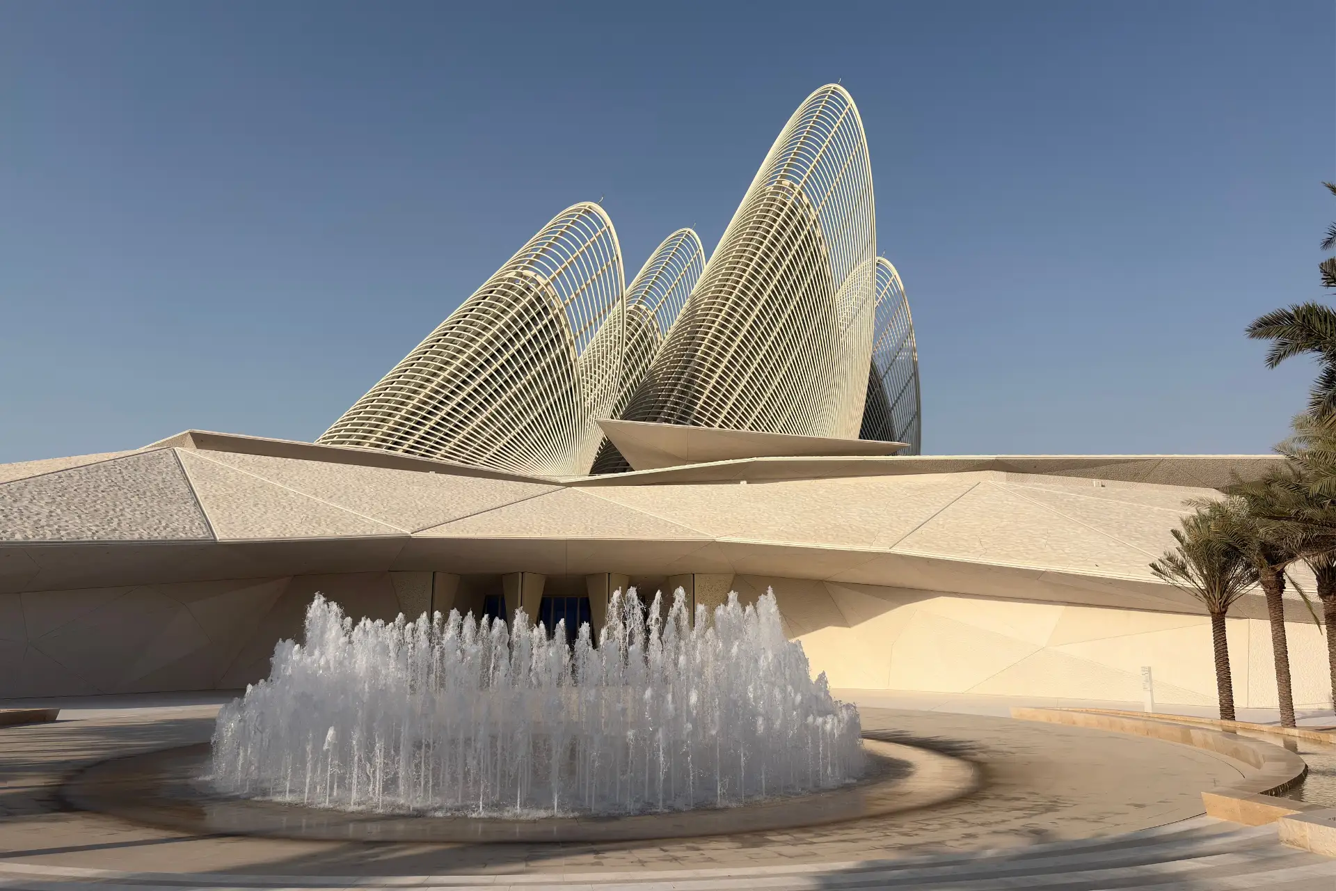 Modern architectural structure with curved metal grids, a white geometric facade, and a round fountain in front, set against a clear blue sky.