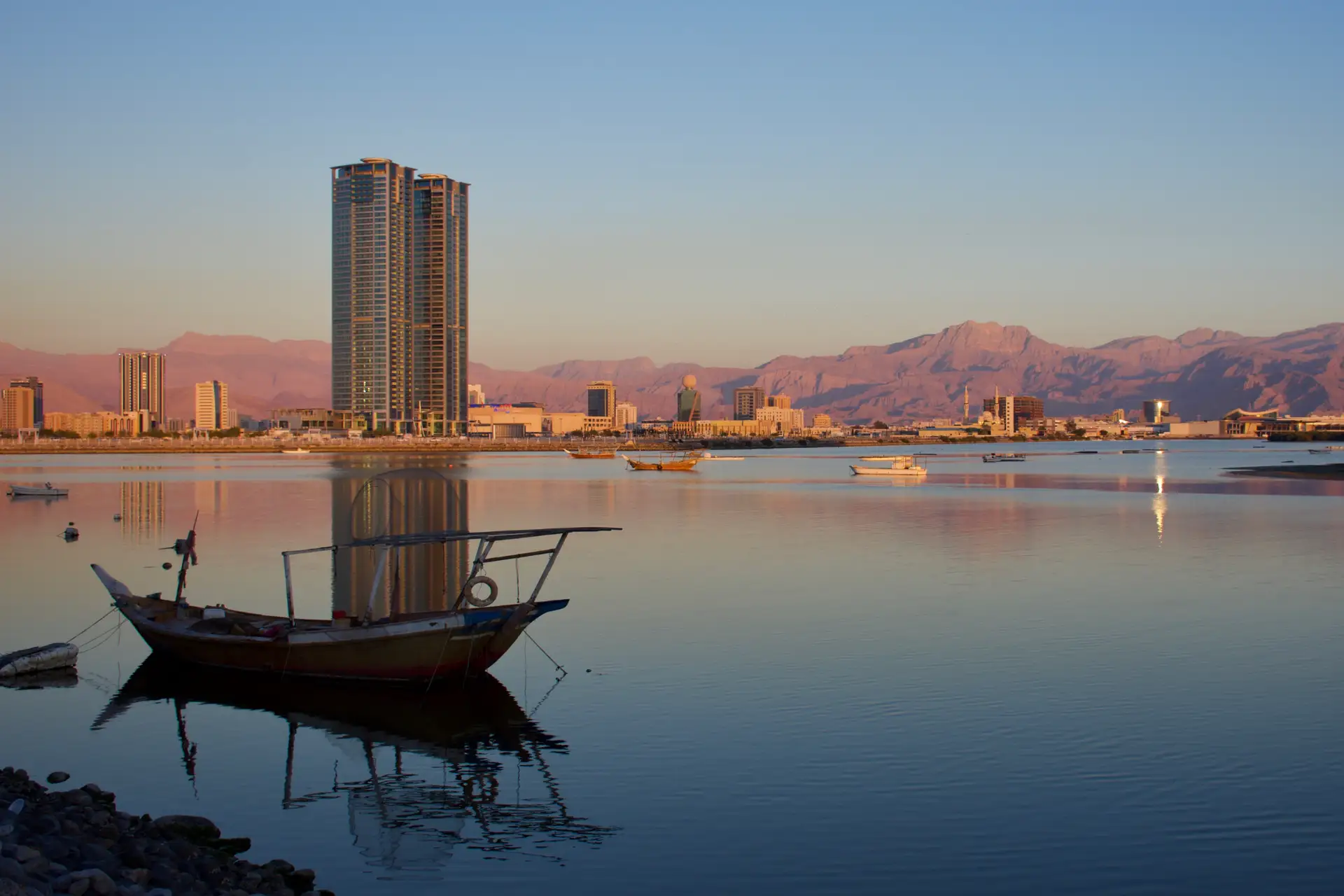 A serene waterfront scene at sunset with a traditional boat, high-rise buildings, and mountains in the background.