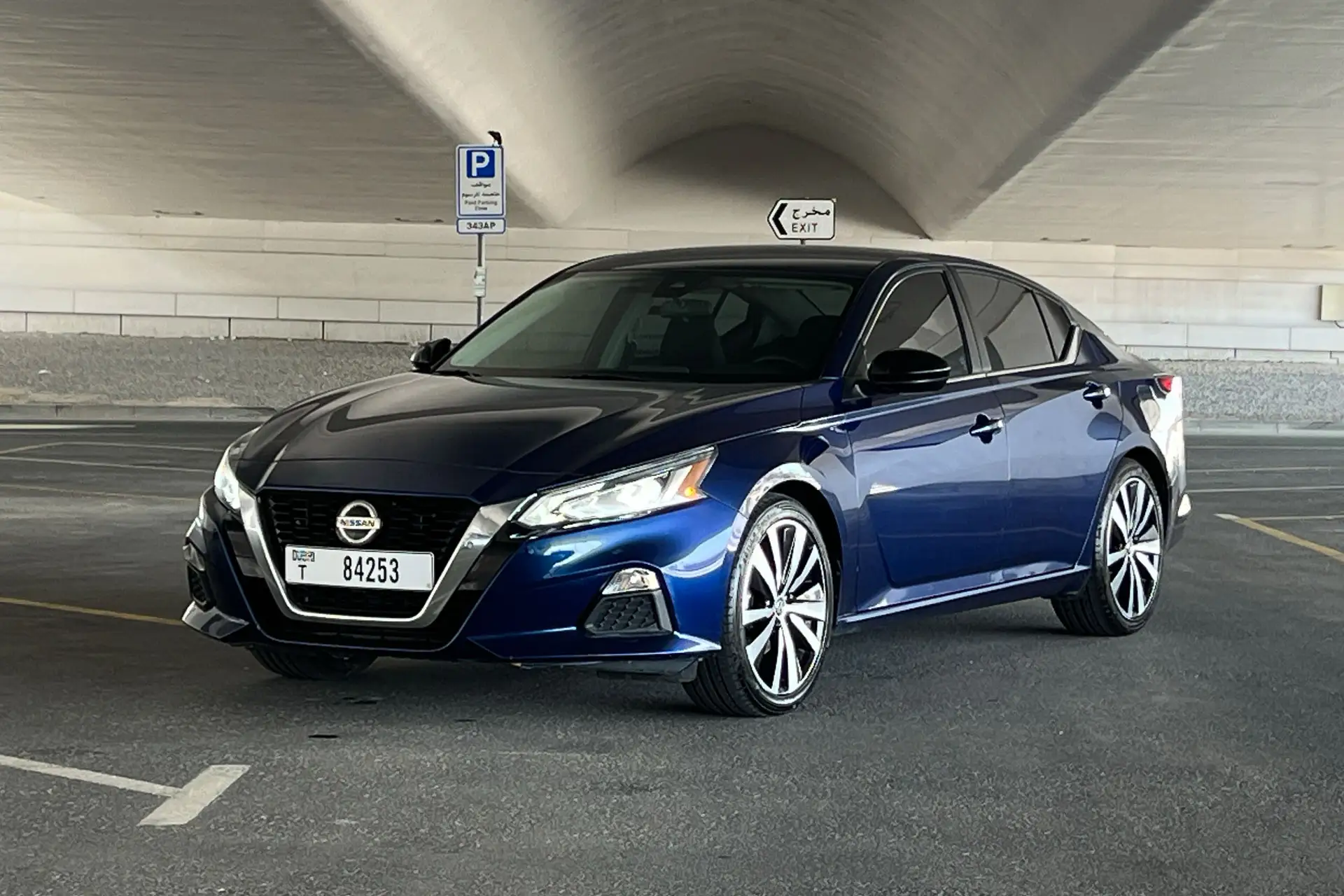 A blue Nissan sedan parked in a shaded area under a bridge, with a "Parking" sign and exit sign visible in the background.