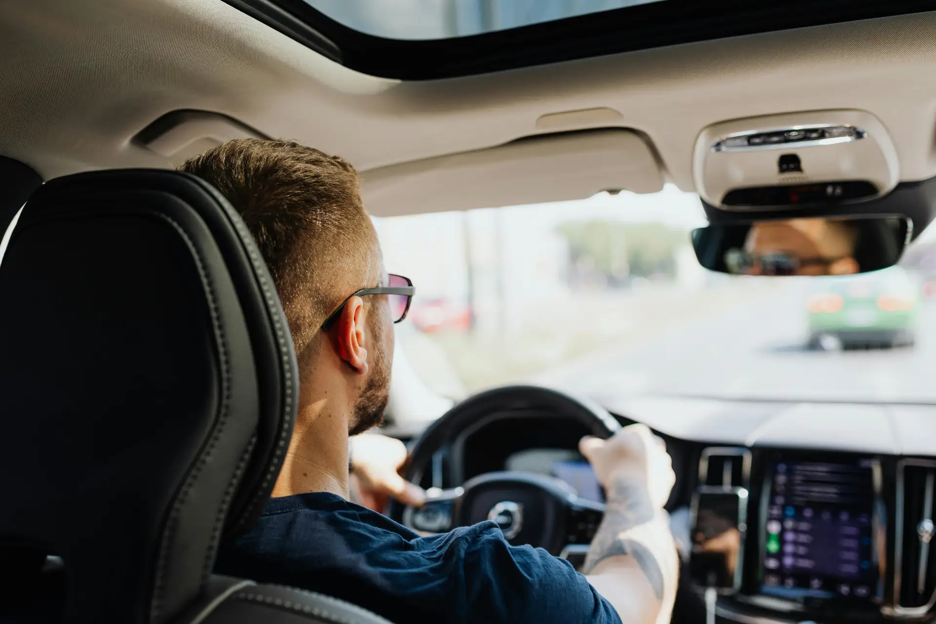 A person with short hair and glasses drives a car with a visible dashboard and rearview mirror, on a sunny day.