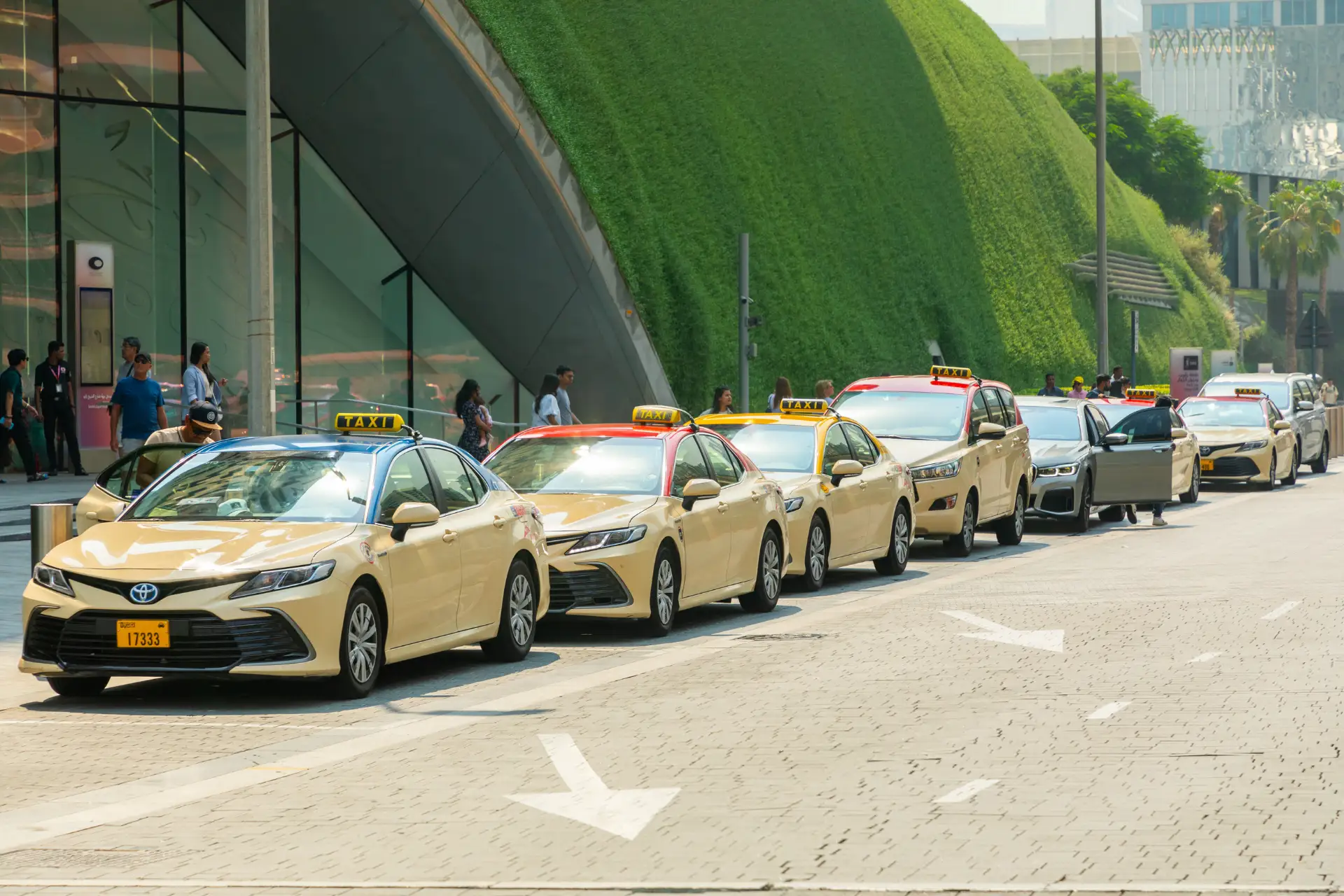 A line of beige taxis parked along a modern building with a green wall, people walking nearby in daylight.