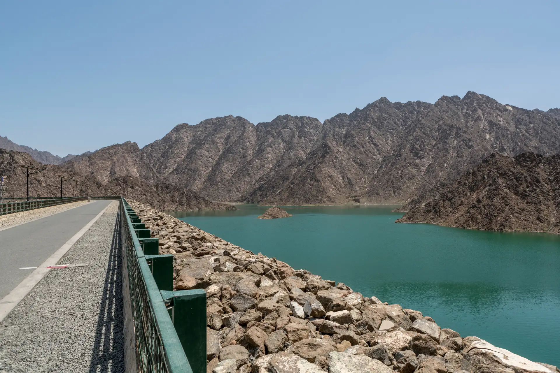 A turquoise reservoir surrounded by rugged mountains, with a paved path and rocky embankment under a clear blue sky.
