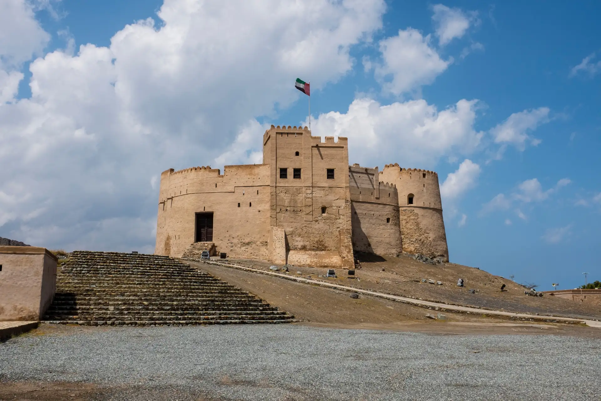 A historic beige fortress with crenellated walls and a flag on top, set against a cloudy blue sky, with a stone path leading up to it.
