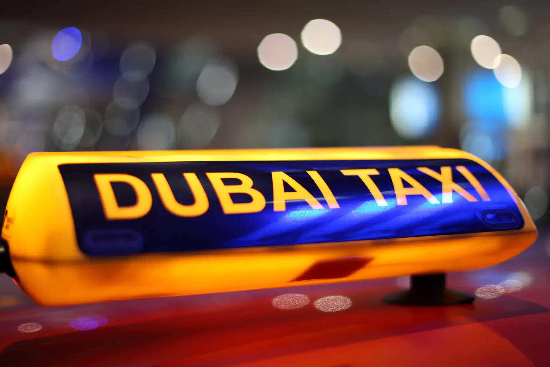 Illuminated "Dubai Taxi" sign on a vehicle roof, with blurred lights in the background.