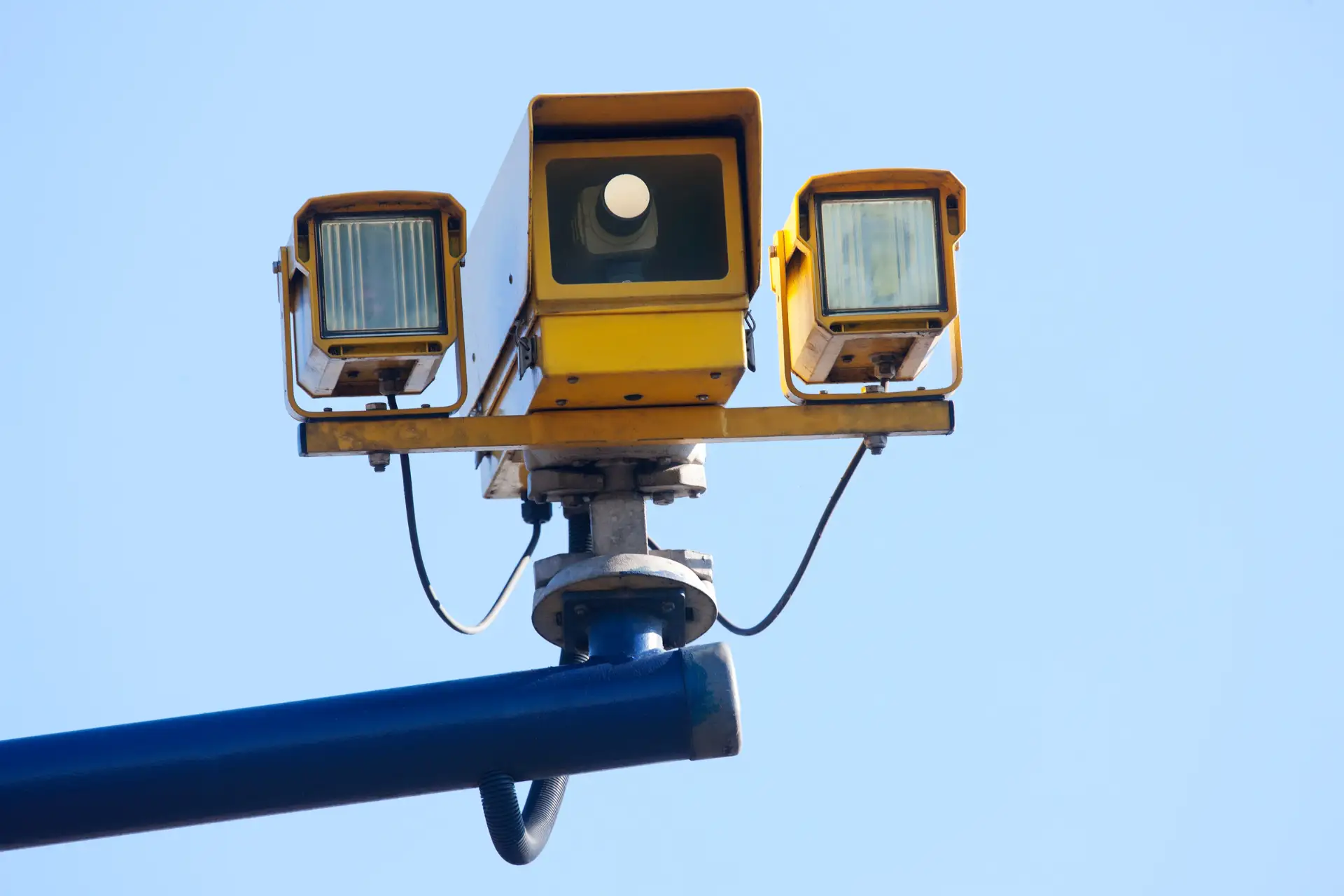 Yellow traffic camera with two lights mounted on a pole against a clear blue sky.