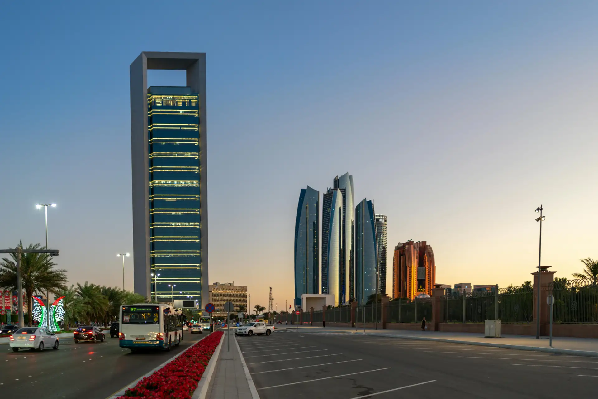 Urban skyline with modern skyscrapers under twilight sky, cars and buses on the street, red flowers lining the road.