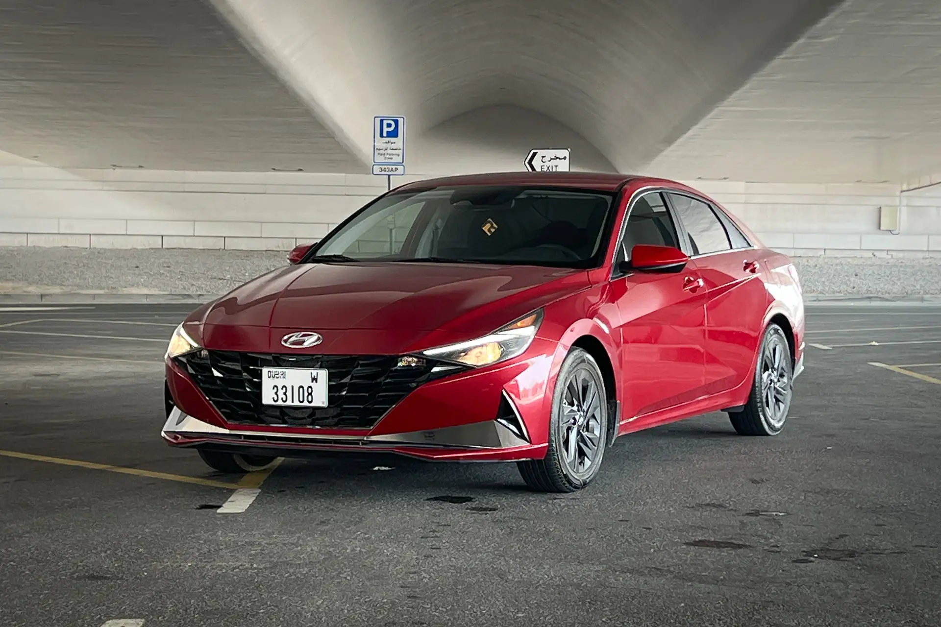 A red sedan labeled "33108" parked in an empty, covered parking area with a visible handicap parking sign in the background.