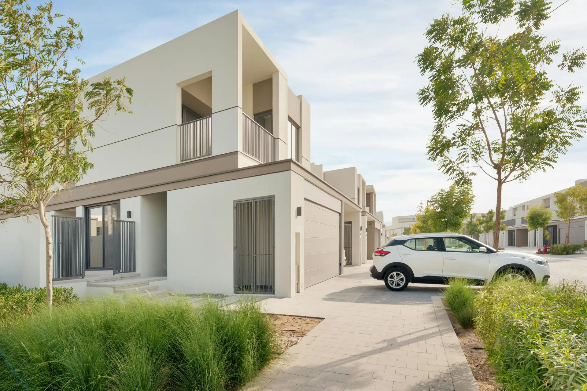 Modern two-story house with a white facade, a driveway with a parked white SUV, and surrounding greenery under a clear sky.