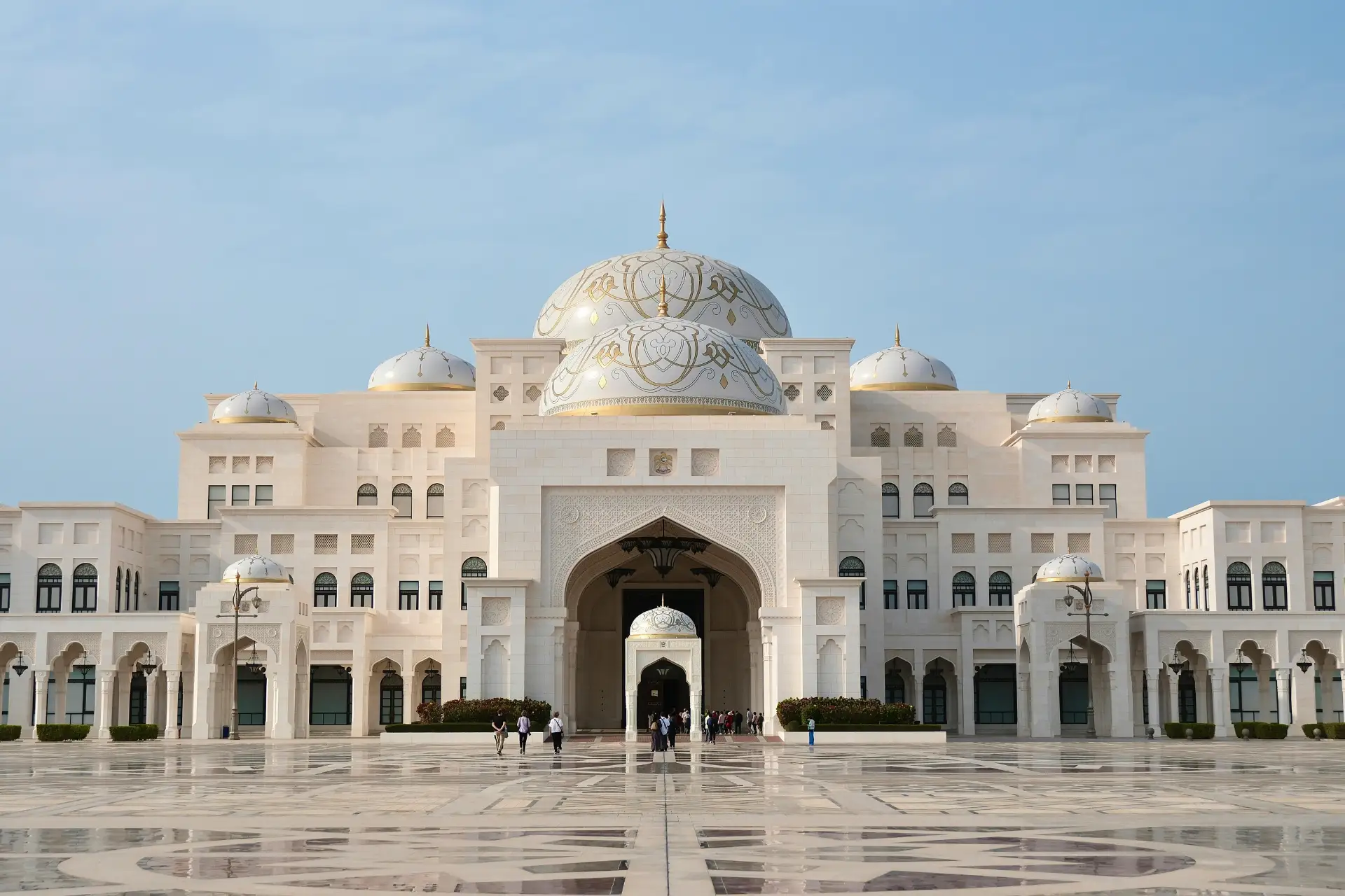 Palace with ornate domes and arches, featuring a large courtyard and a group of people walking towards the entrance under a clear blue sky.