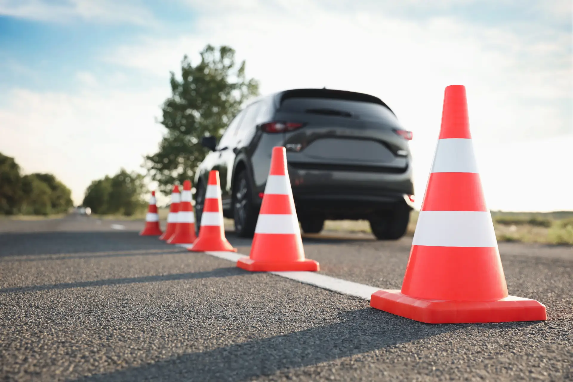 Orange traffic cones line a road with a parked car and trees in the background.