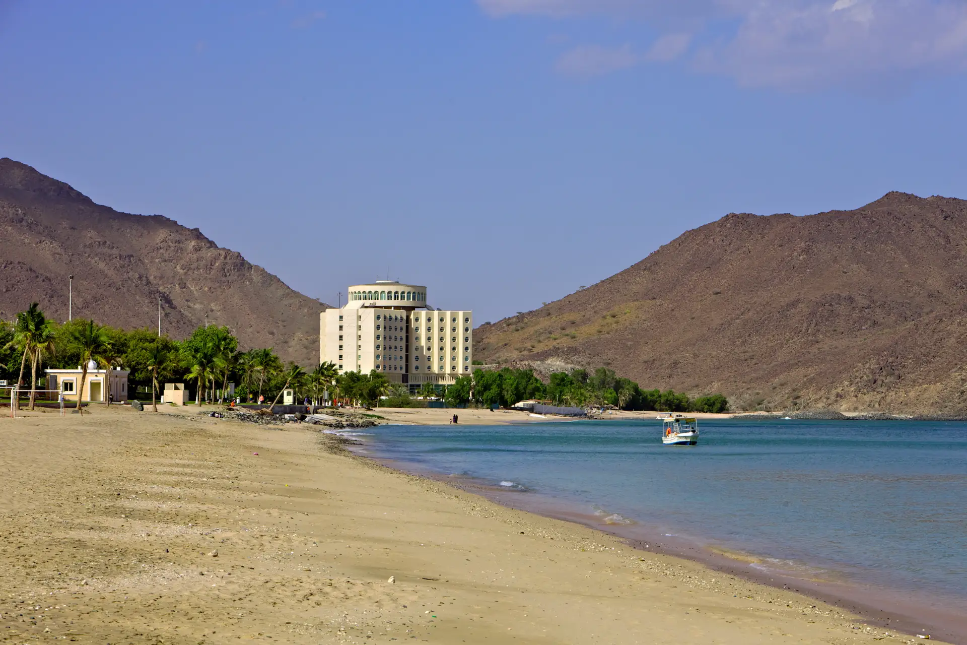A large building with arches stands near a beach and palm trees, framed by arid mountains with a small boat in the turquoise water.
