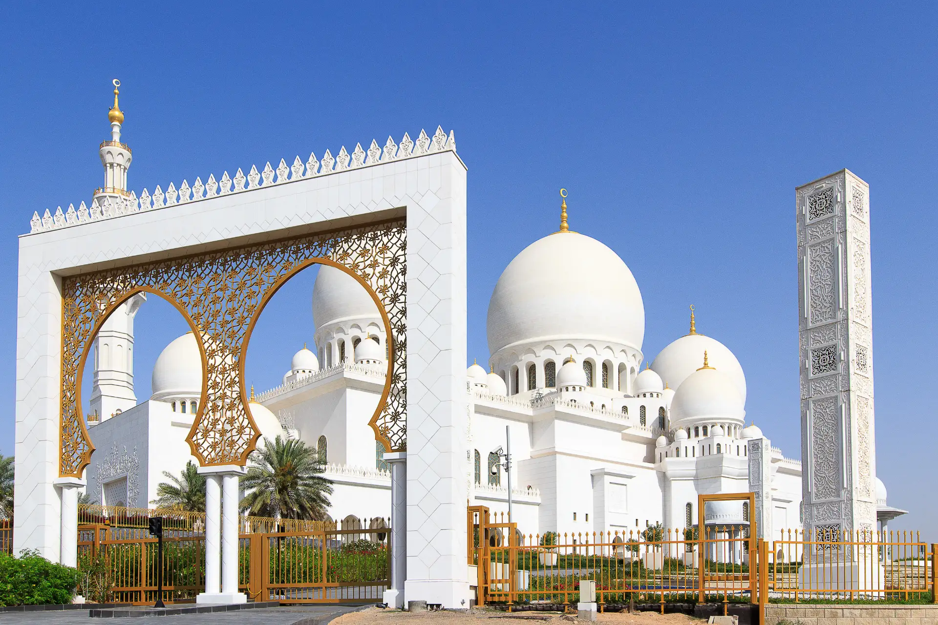 White mosque with large domes, ornate arch, and minaret against a clear blue sky.