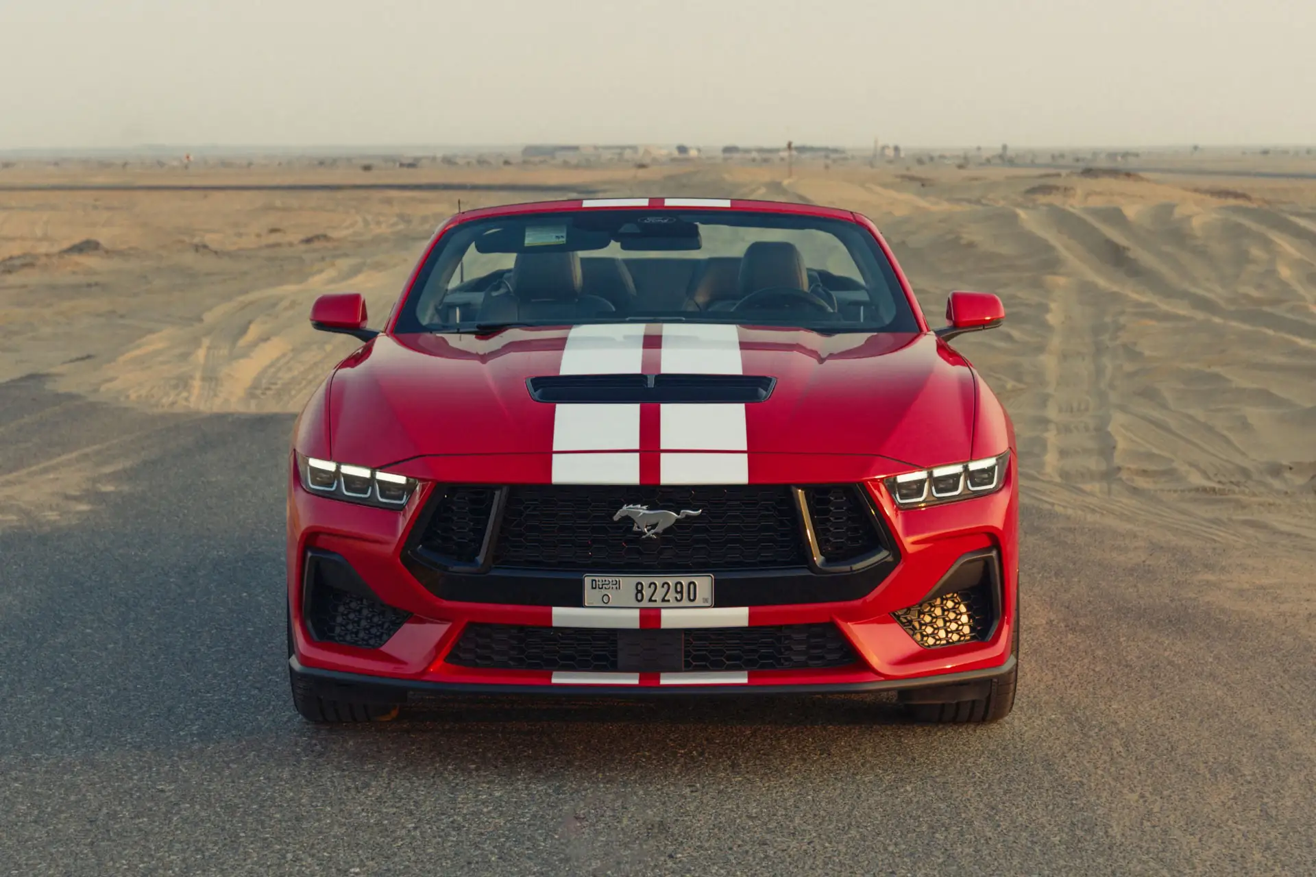Red convertible sports car with white racing stripes on a desert road. Mustang logo on the grille. Desert landscape in the background.