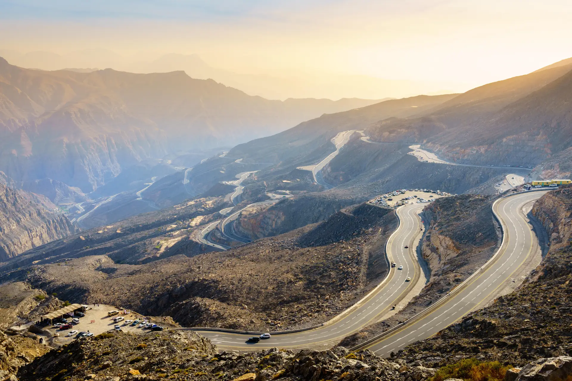 Winding mountain road with several cars, set against a rugged landscape and a hazy sky at sunset.