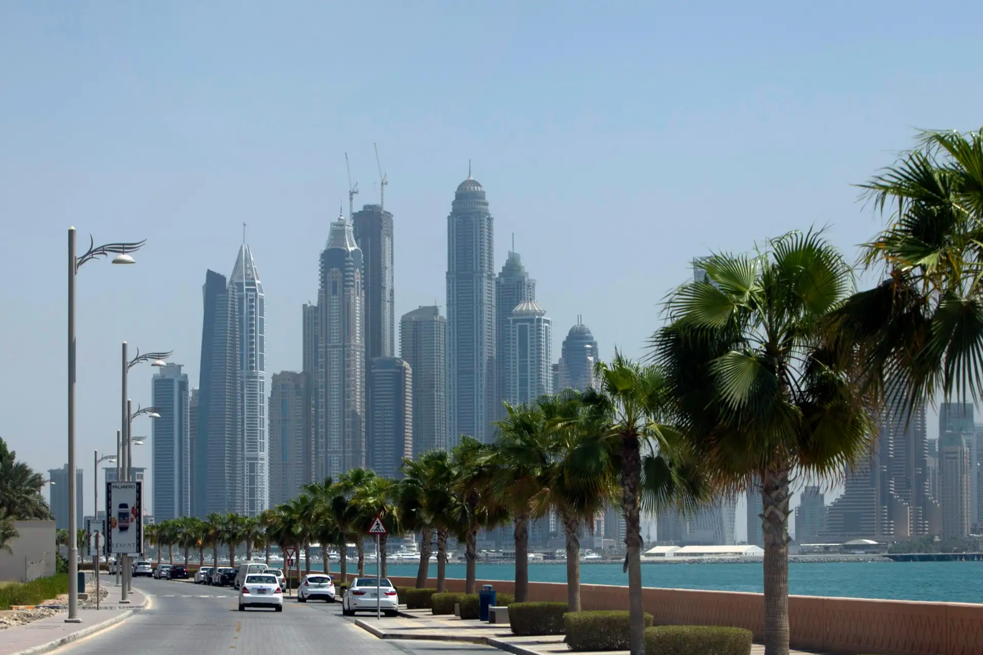 Palm tree-lined street with cars driving, leading towards a skyline of modern skyscrapers near a waterfront under a clear blue sky.